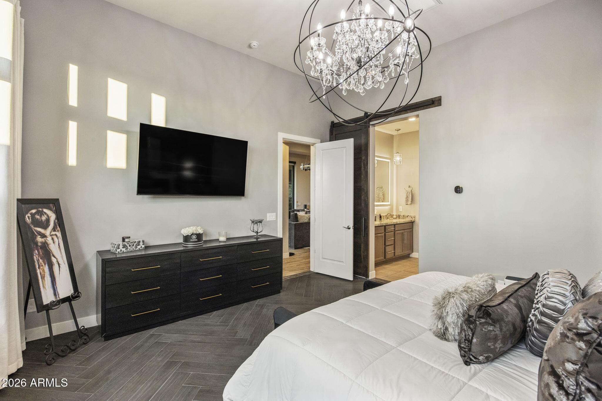 Bedroom with a white bed, crystal chandelier, wall-mounted TV, and open doorway to the bathroom.