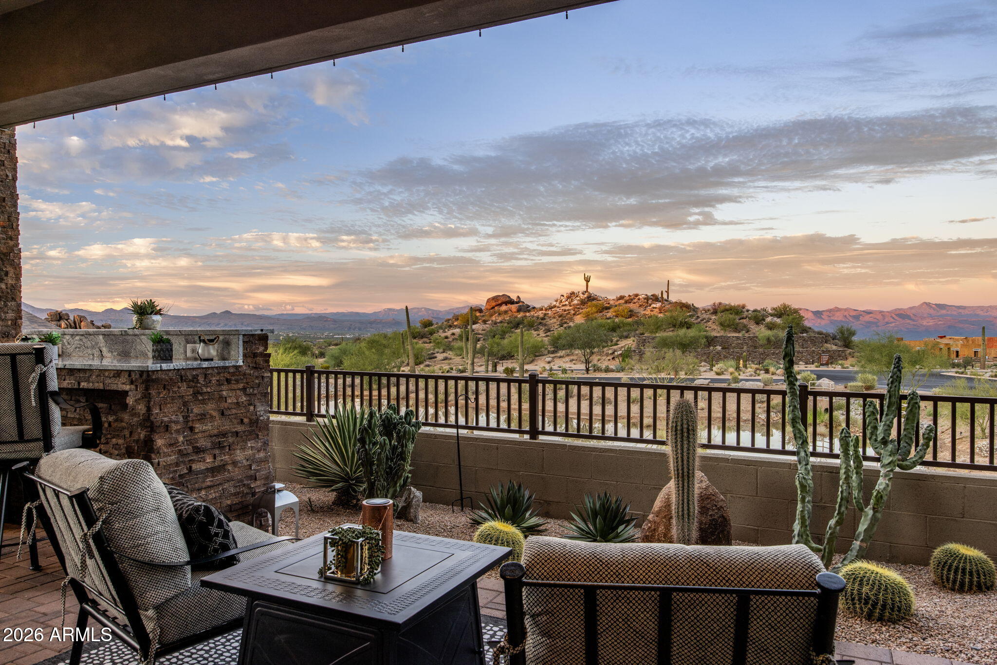 Covered patio with cushioned seating overlooks a cactus-dotted desert hillside and distant mountains at sunset.