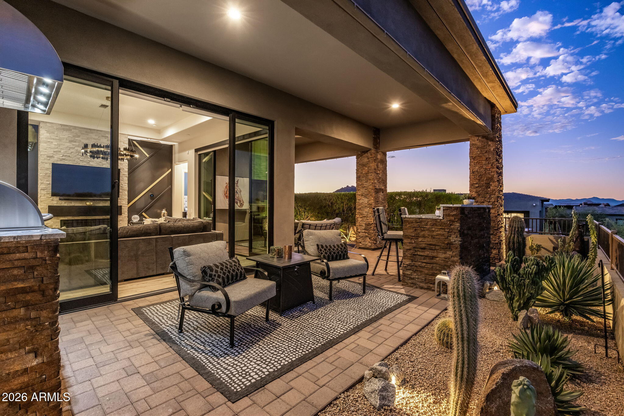 Covered patio with cushioned seating, a stone outdoor bar, desert plants, and distant mountains at dusk.