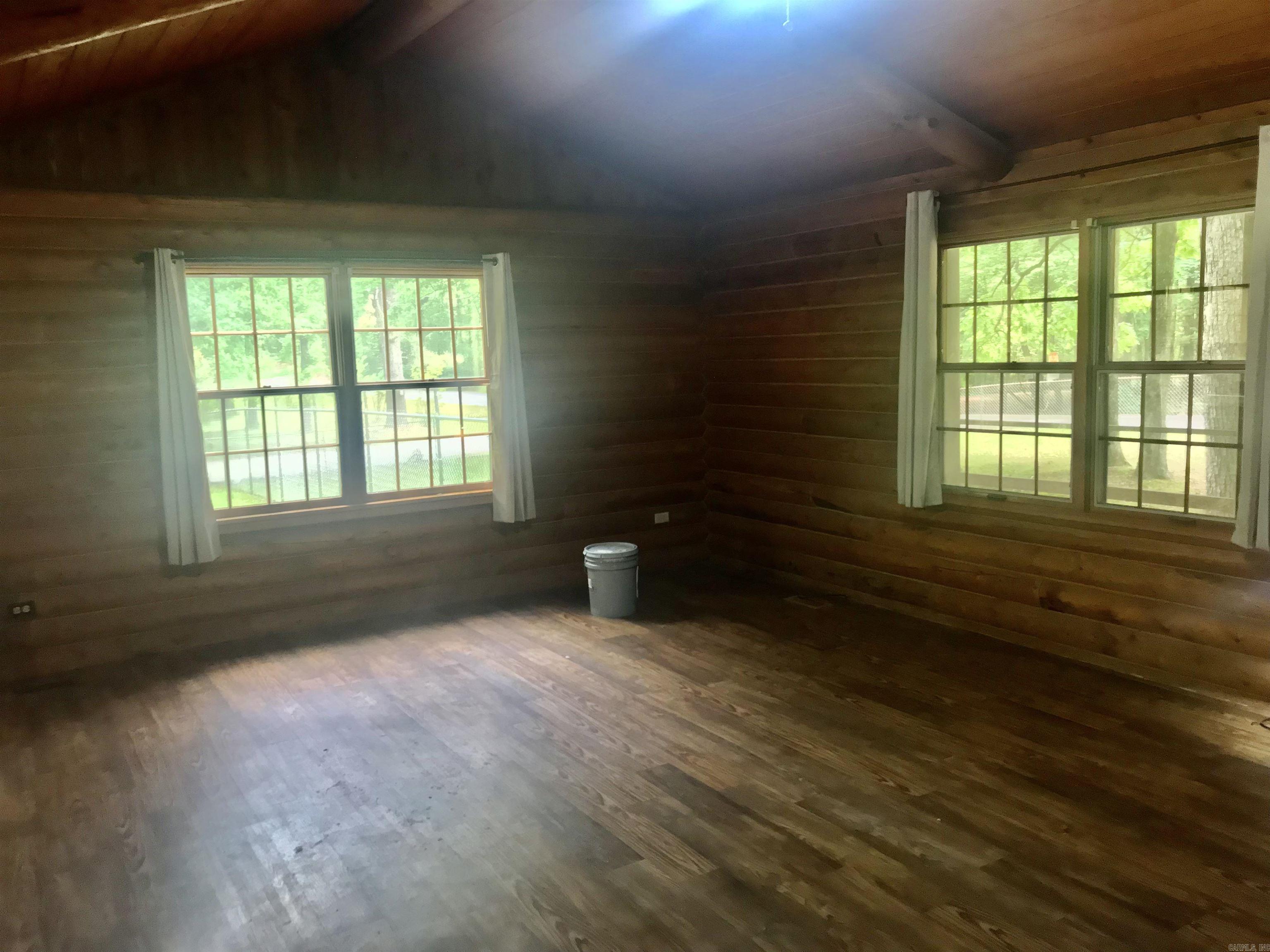 Vacant wood-paneled room with a worn wood floor and two sets of windows overlooking trees.