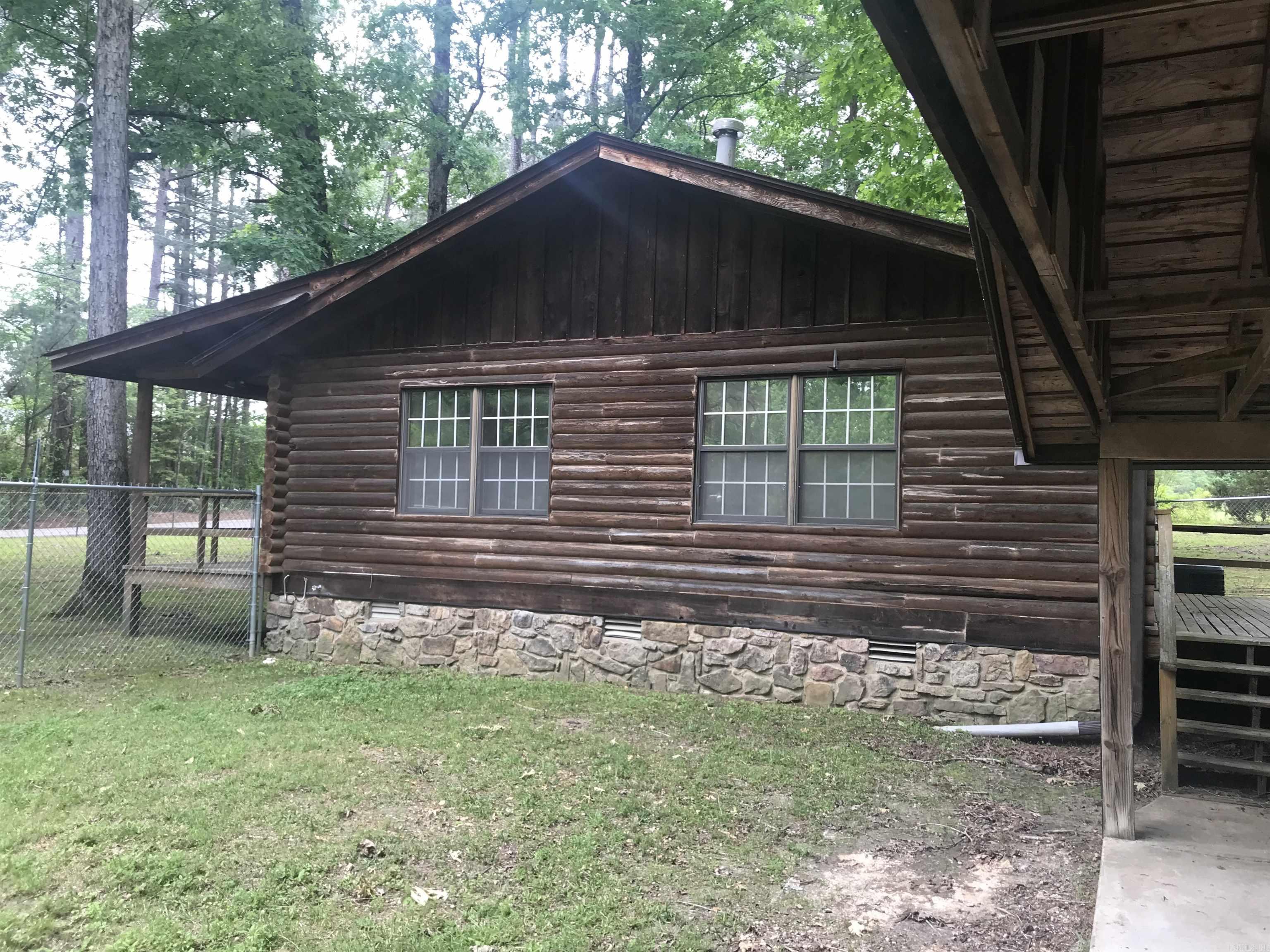 Log cabin side with stone foundation and white-grid windows, beneath trees beside a covered deck.