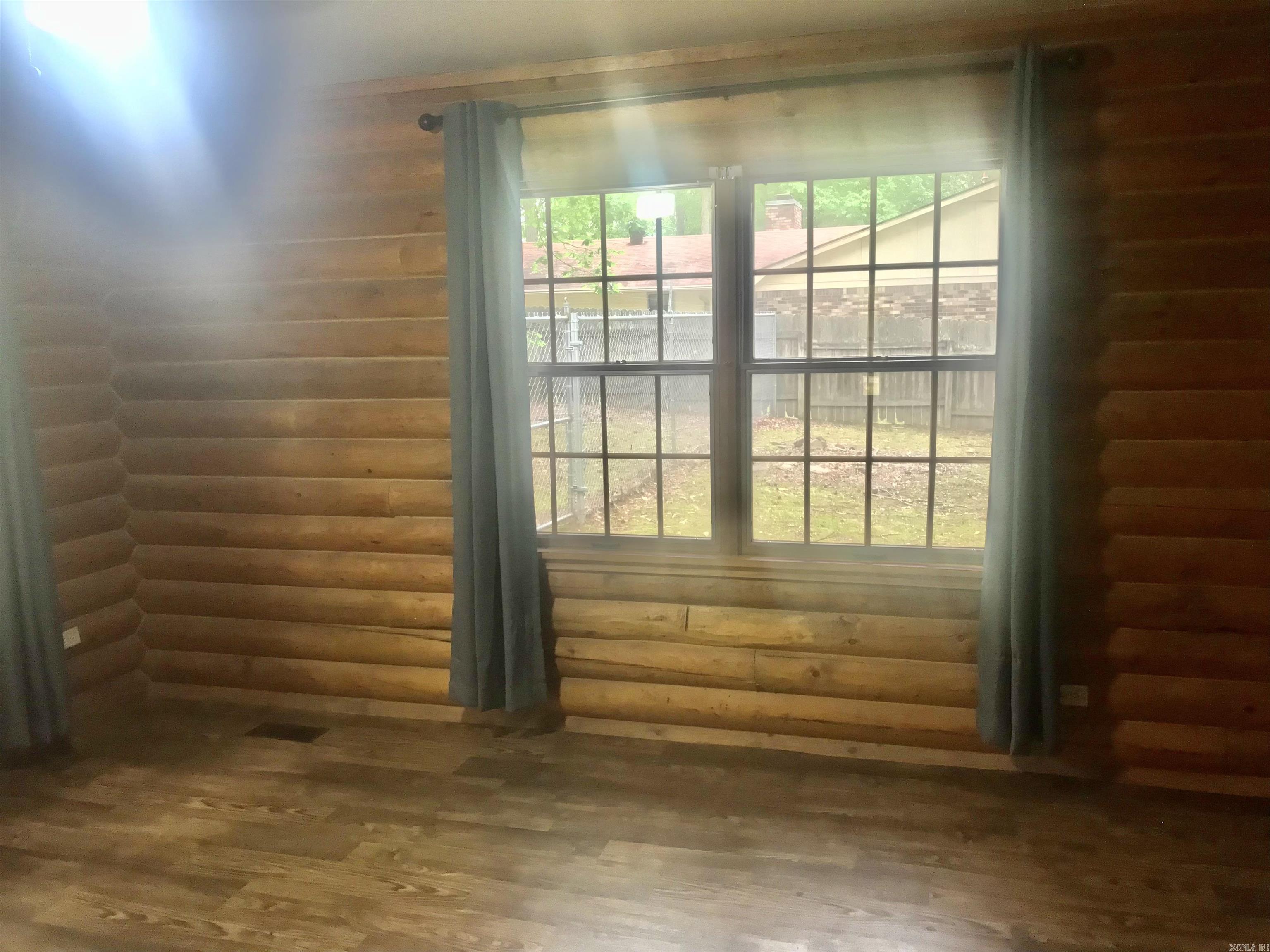 Empty log-walled room with wood floors and a large curtained window.