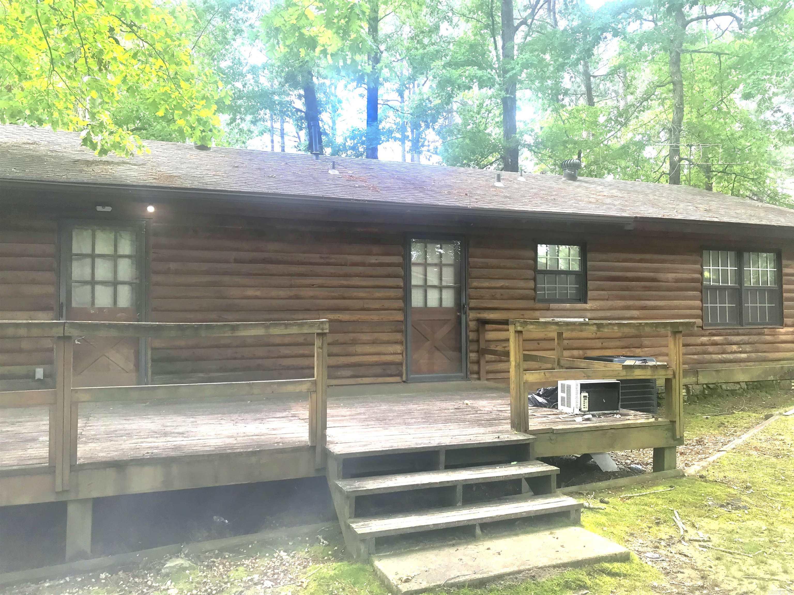 Log-sided home with a raised wooden deck and steps beneath mature trees.
