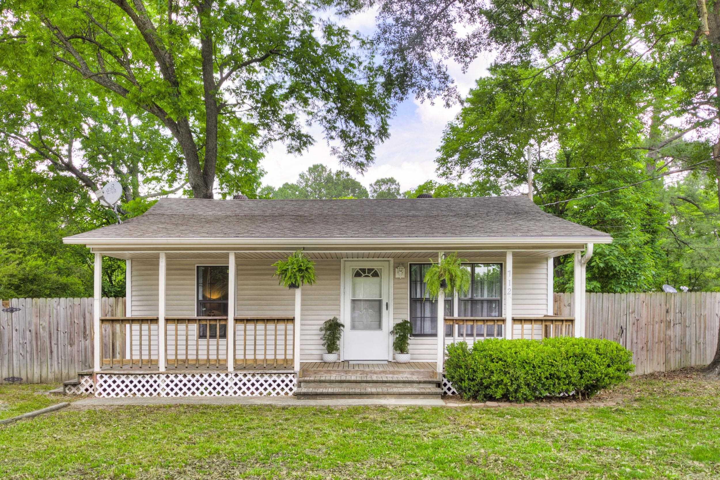 Single-story house with a covered porch, hanging ferns, and a fenced yard beneath mature trees.