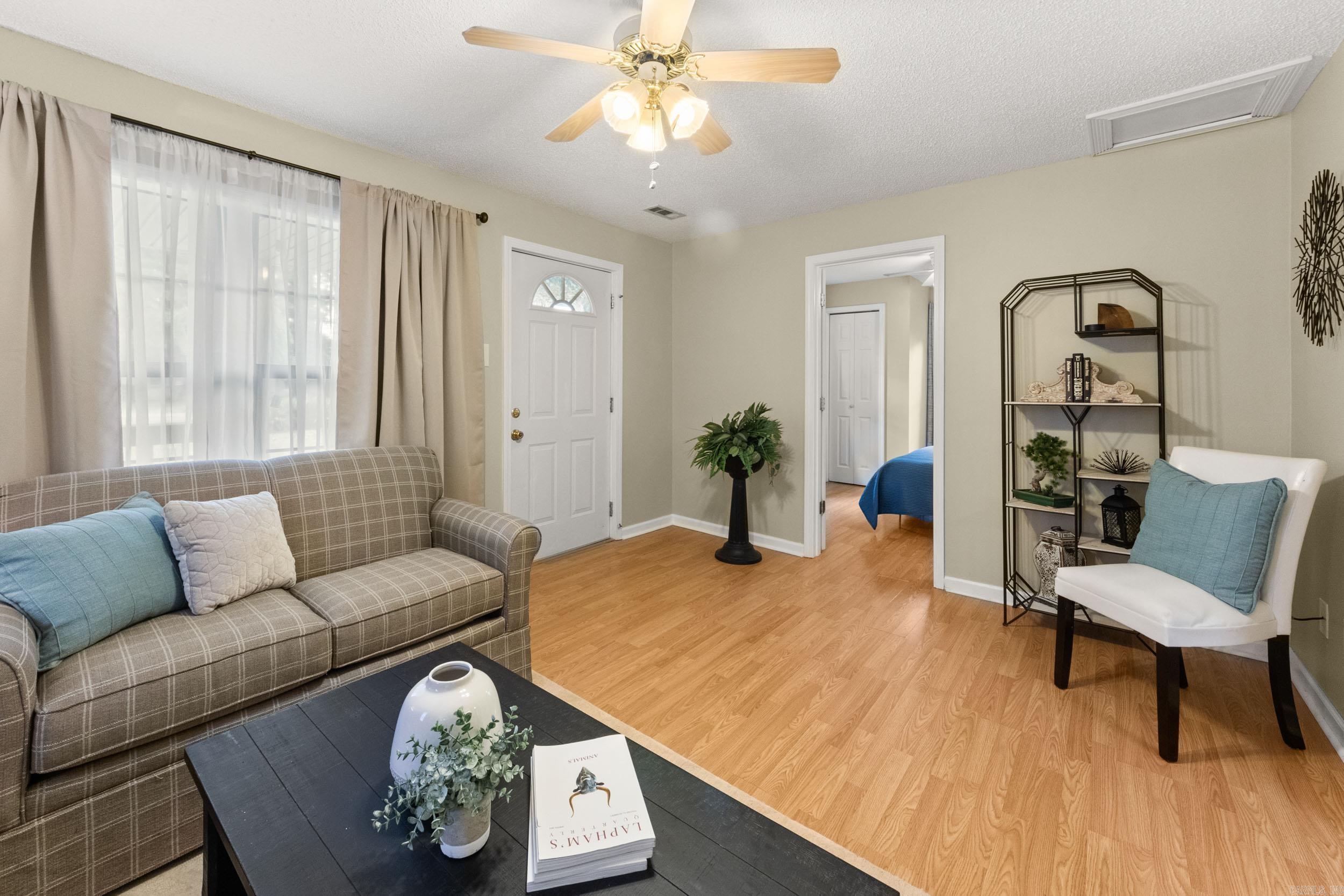 Living room with a plaid sofa, white accent chair, wood-look floors, and a view into the bedroom.