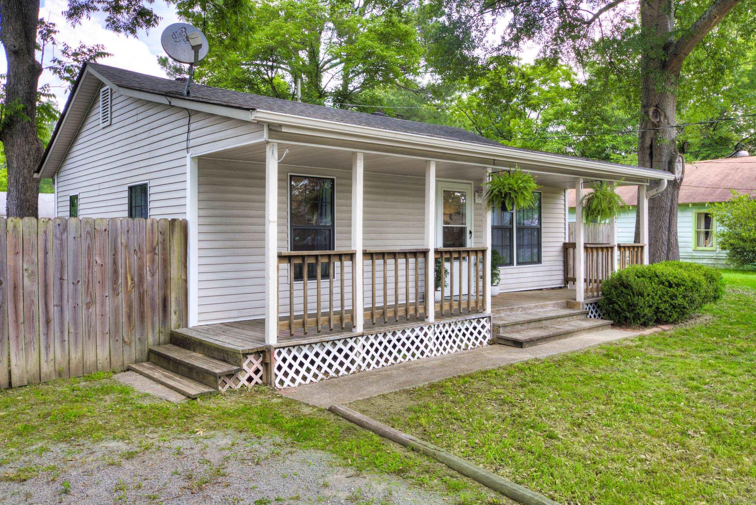 Single-story home with a long covered porch, hanging ferns, and wooden railings beneath mature trees.