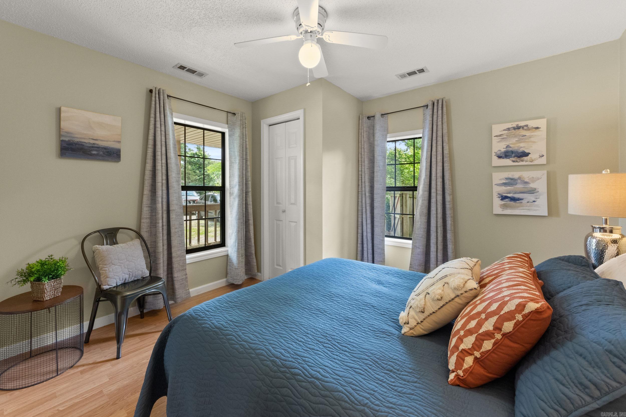 Staged bedroom with a blue quilted bed, two curtained windows, and a ceiling fan.