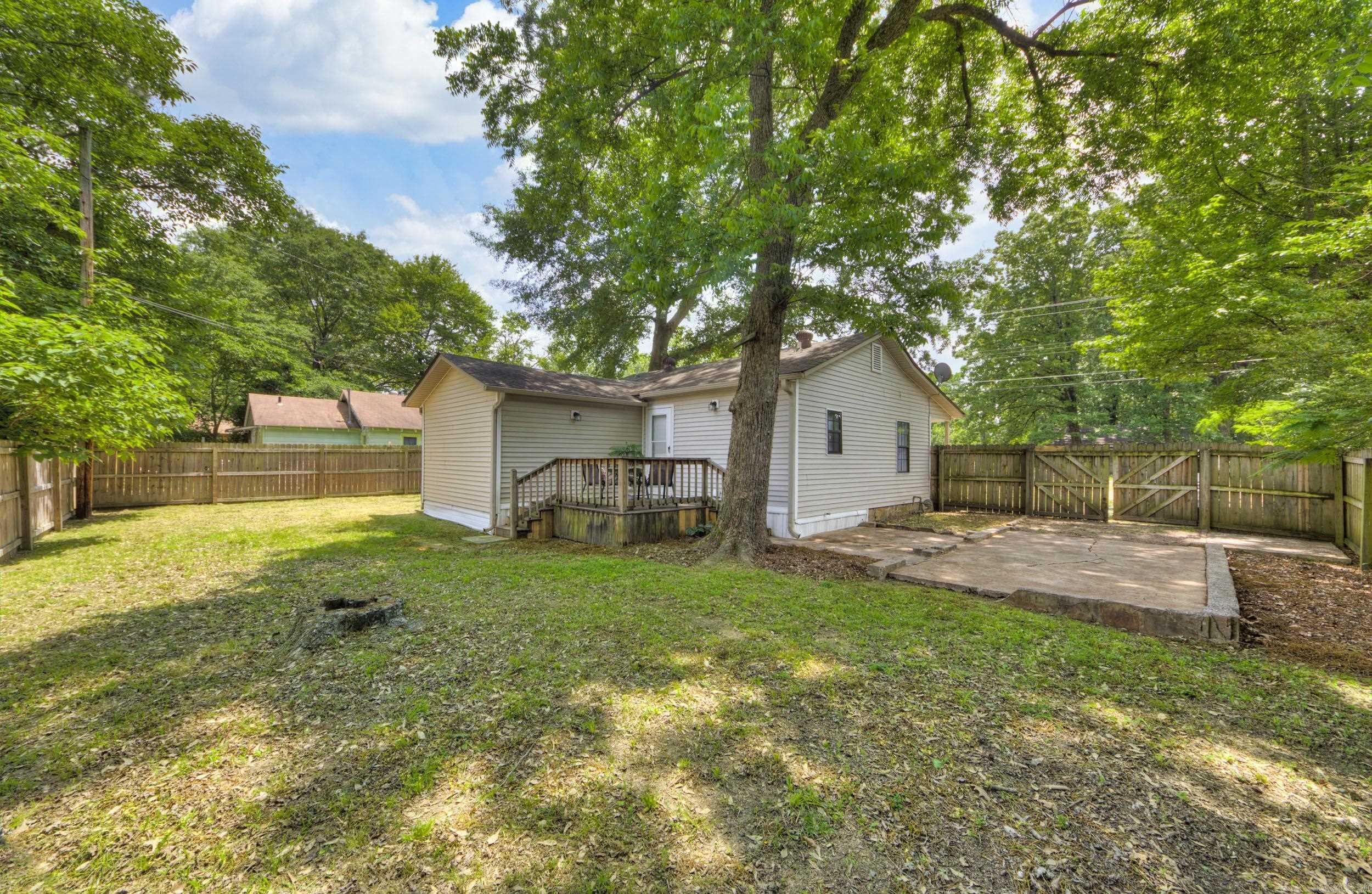 Home’s rear exterior with a small deck, concrete patio, fenced yard, and large shade tree.