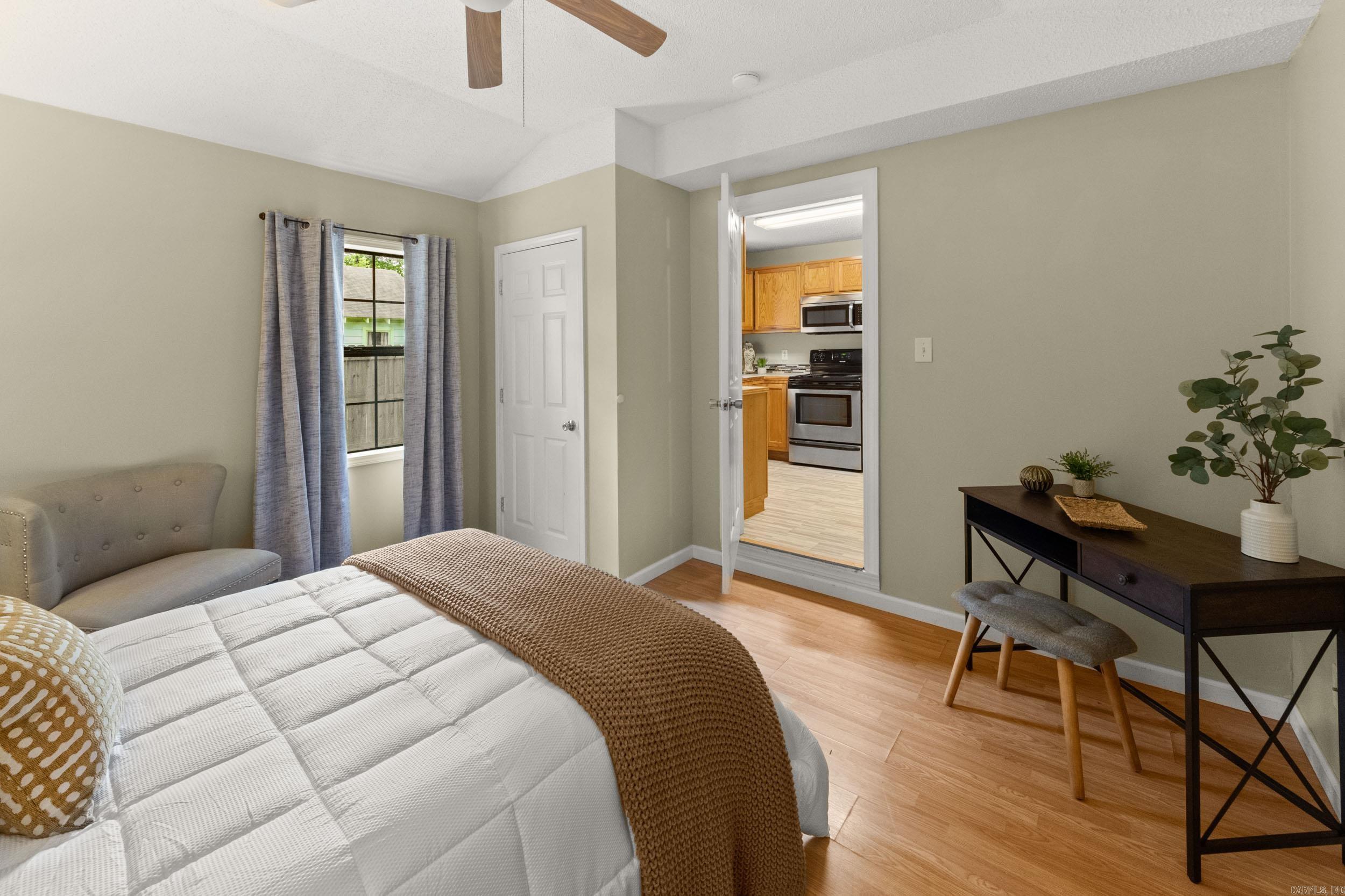 Furnished bedroom with a white bed, brown throw, curtained window, and desk beside an open kitchen doorway.