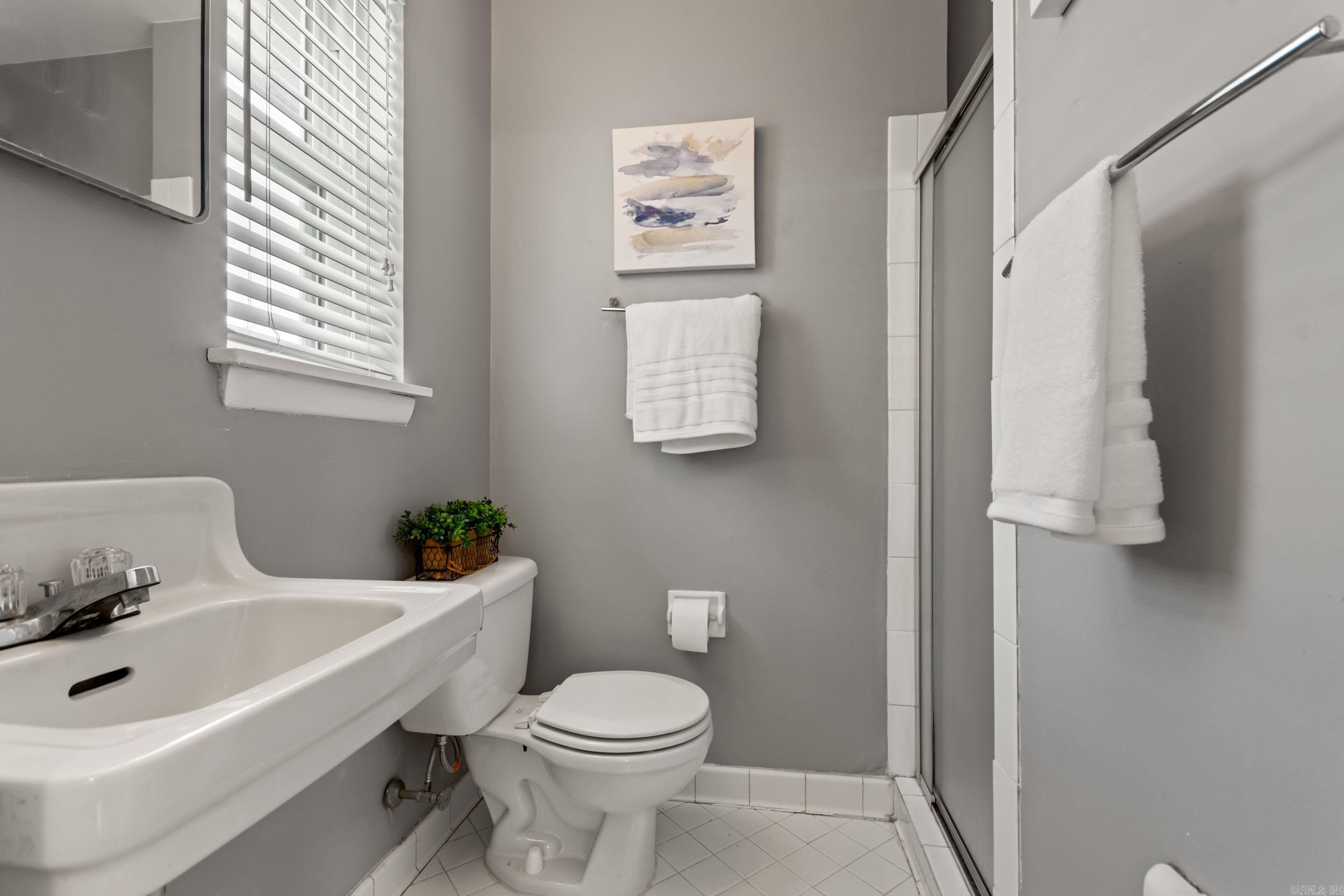Bathroom with a white sink and toilet, glass shower enclosure, and window with blinds.