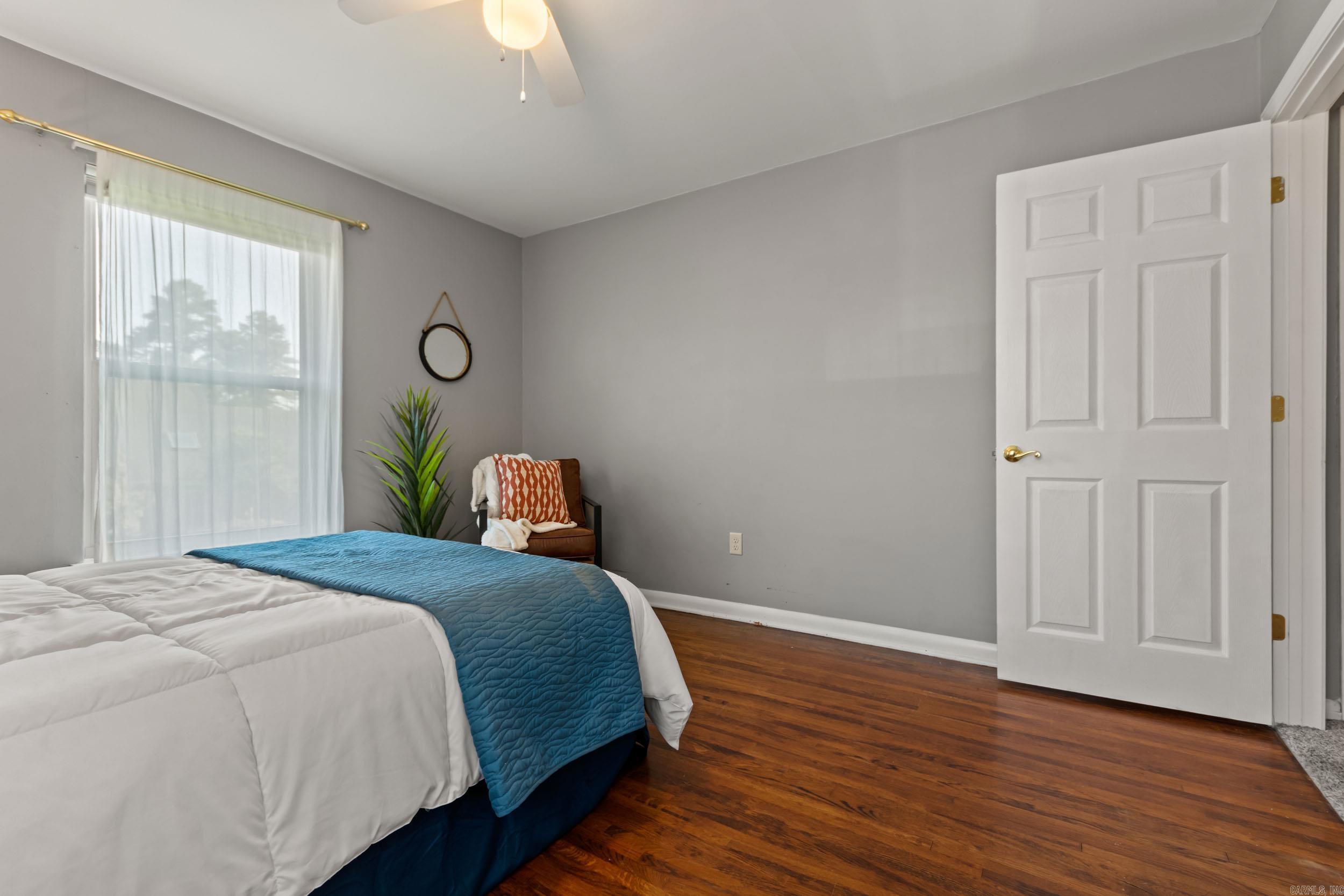 Furnished bedroom with a blue quilt on the bed, hardwood floors, and a large curtained window.
