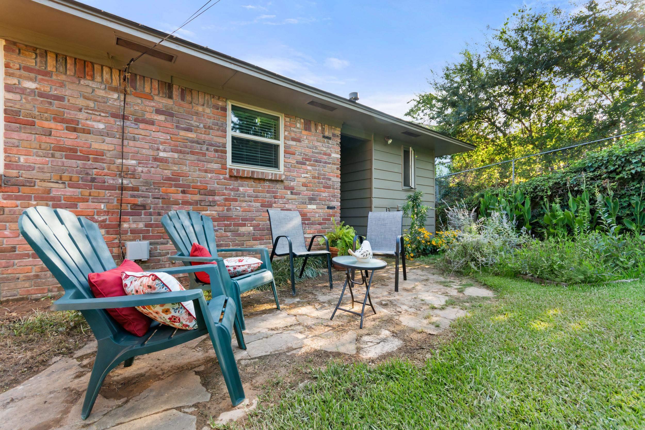 Backyard patio with colorful chairs, stone paving, and a lush garden beside a brick house.