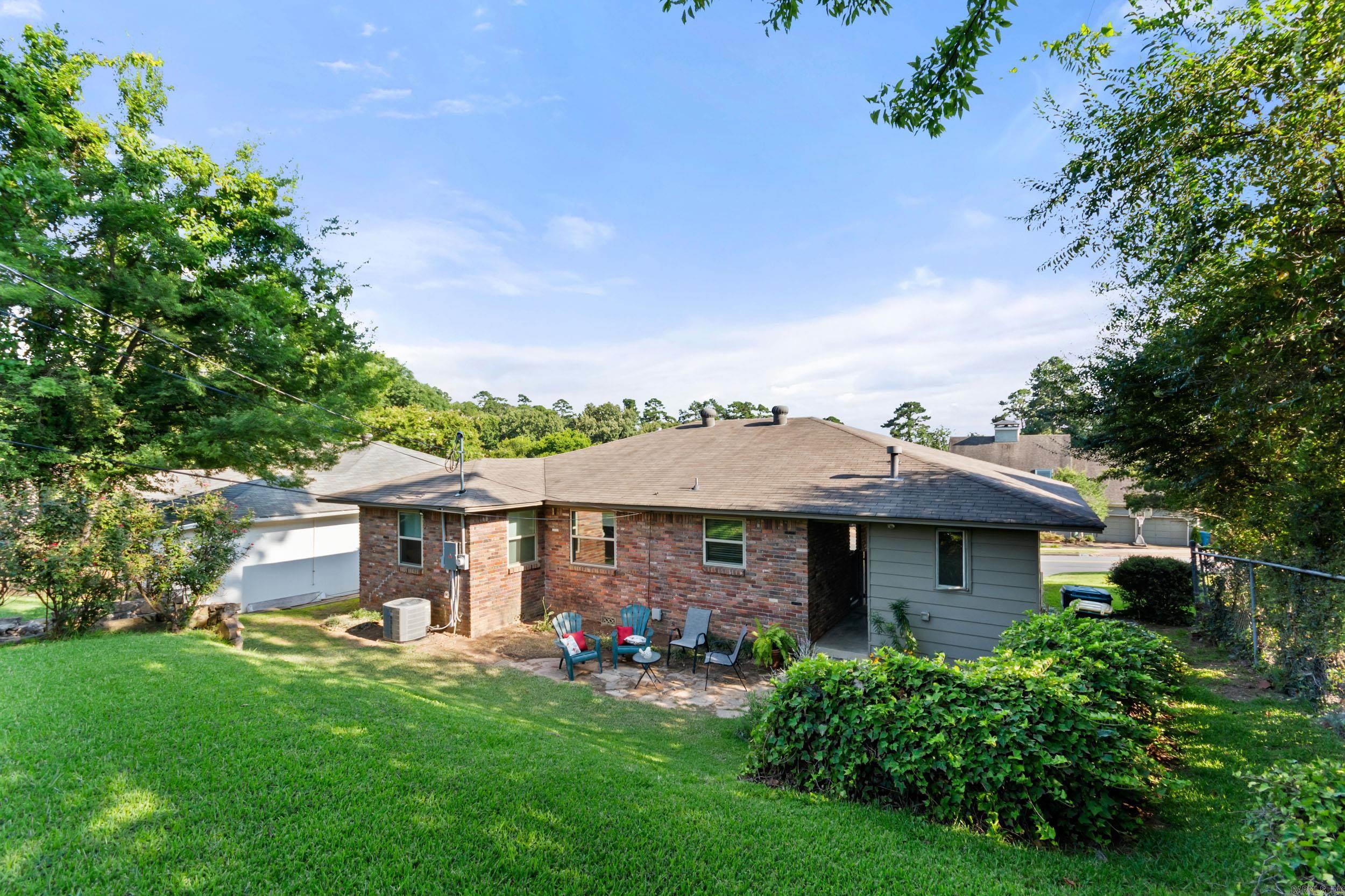 Rear of a brick house with a patio, outdoor chairs, and a sloping lawn beneath mature trees.