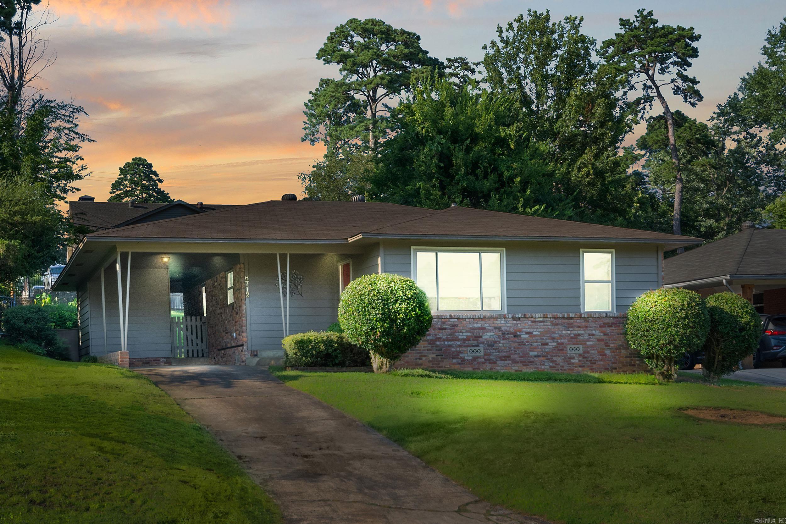 Single-story gray-and-brick home with a covered entry, trimmed shrubs, and a driveway at sunset.