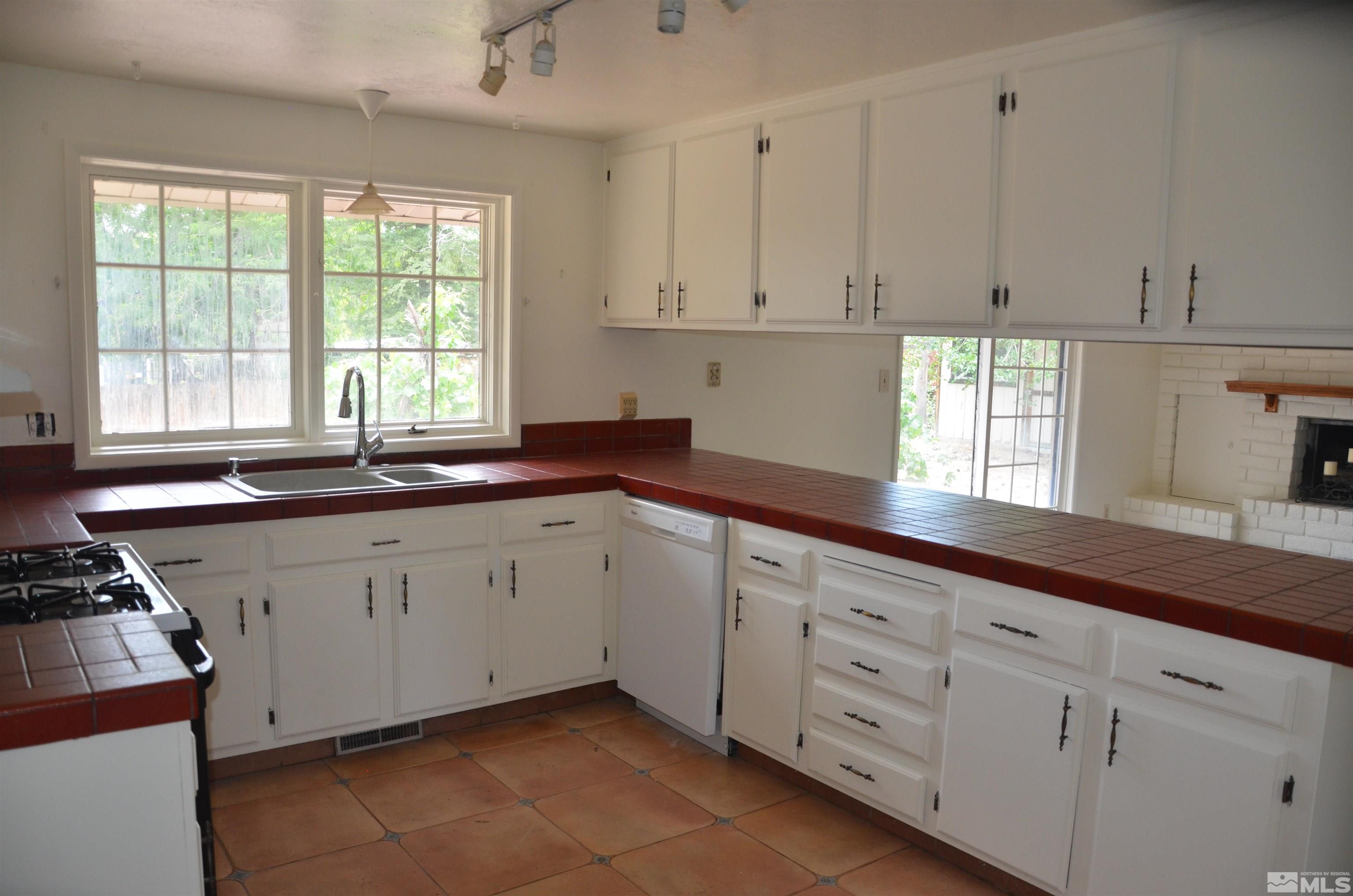 Kitchen with white cabinets, red tile counters, terracotta floors, and large windows beside a brick fireplace.