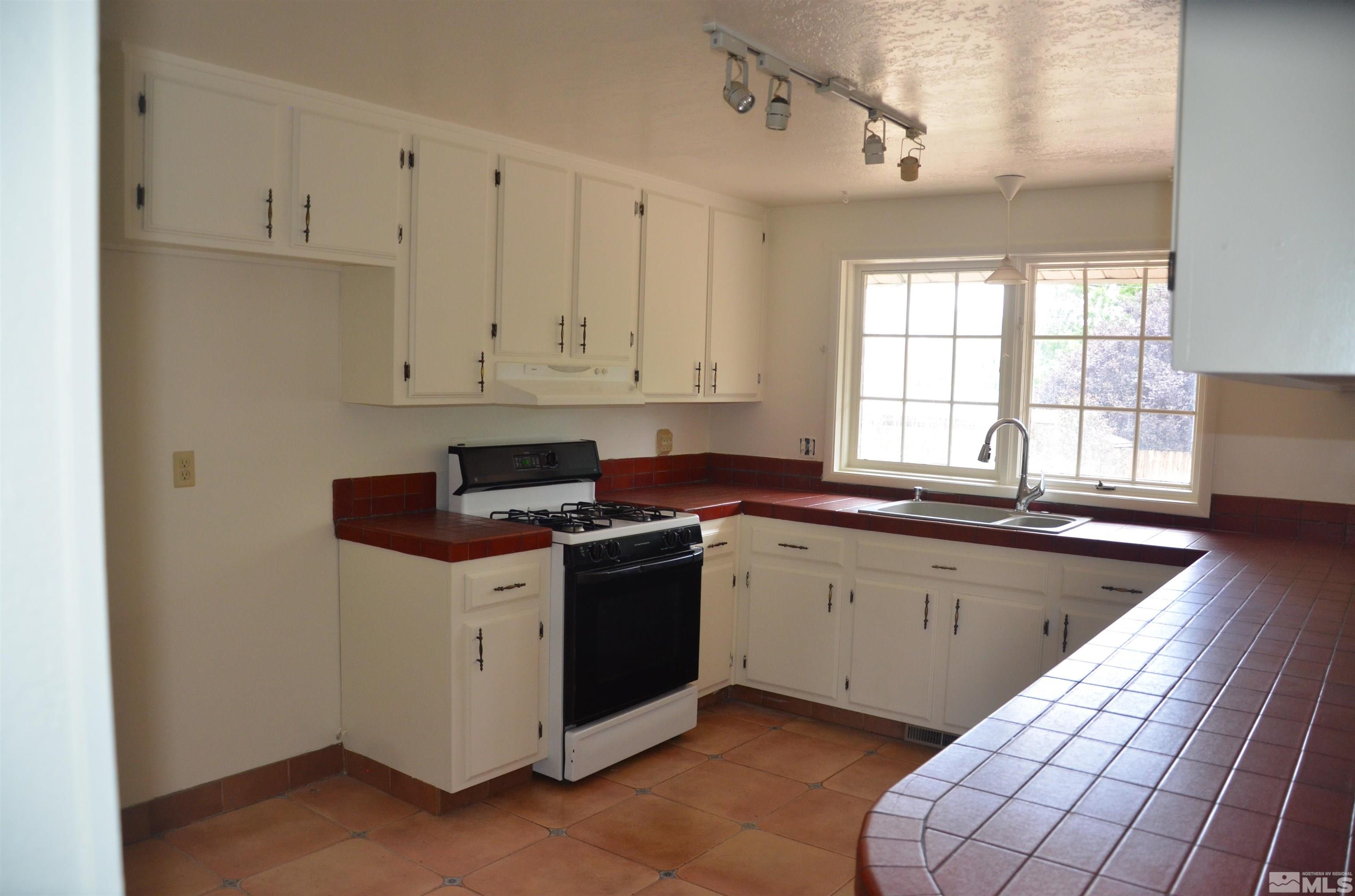 Kitchen with white cabinets, red tile counters, a gas range, and a double sink beneath large windows.