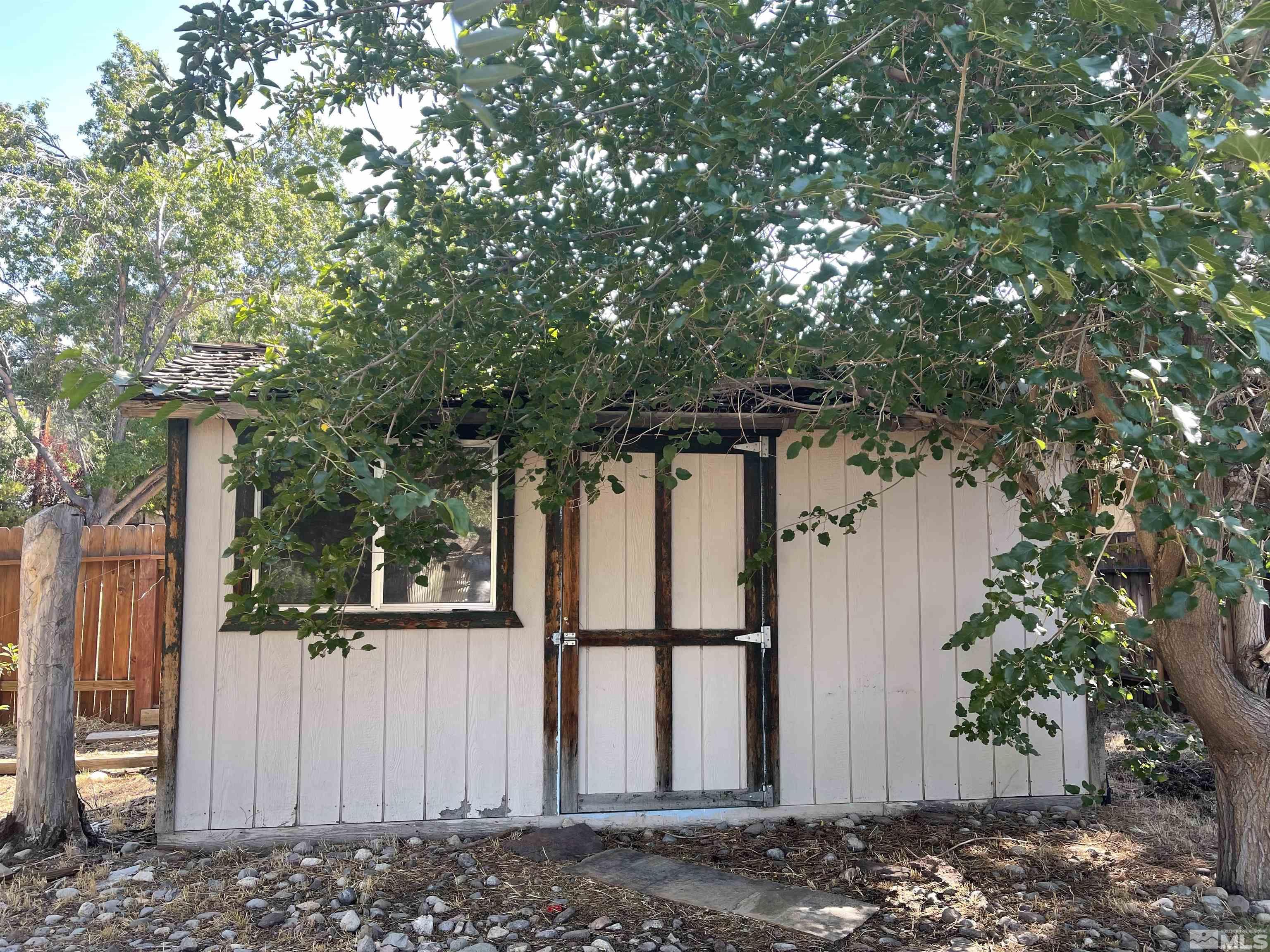 White outbuilding with dark timber trim and double doors beneath overhanging trees.