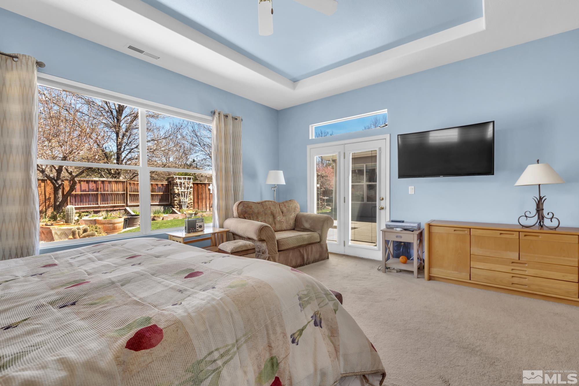 Bedroom with pale blue walls, a tray ceiling, and large windows overlooking a fenced garden.