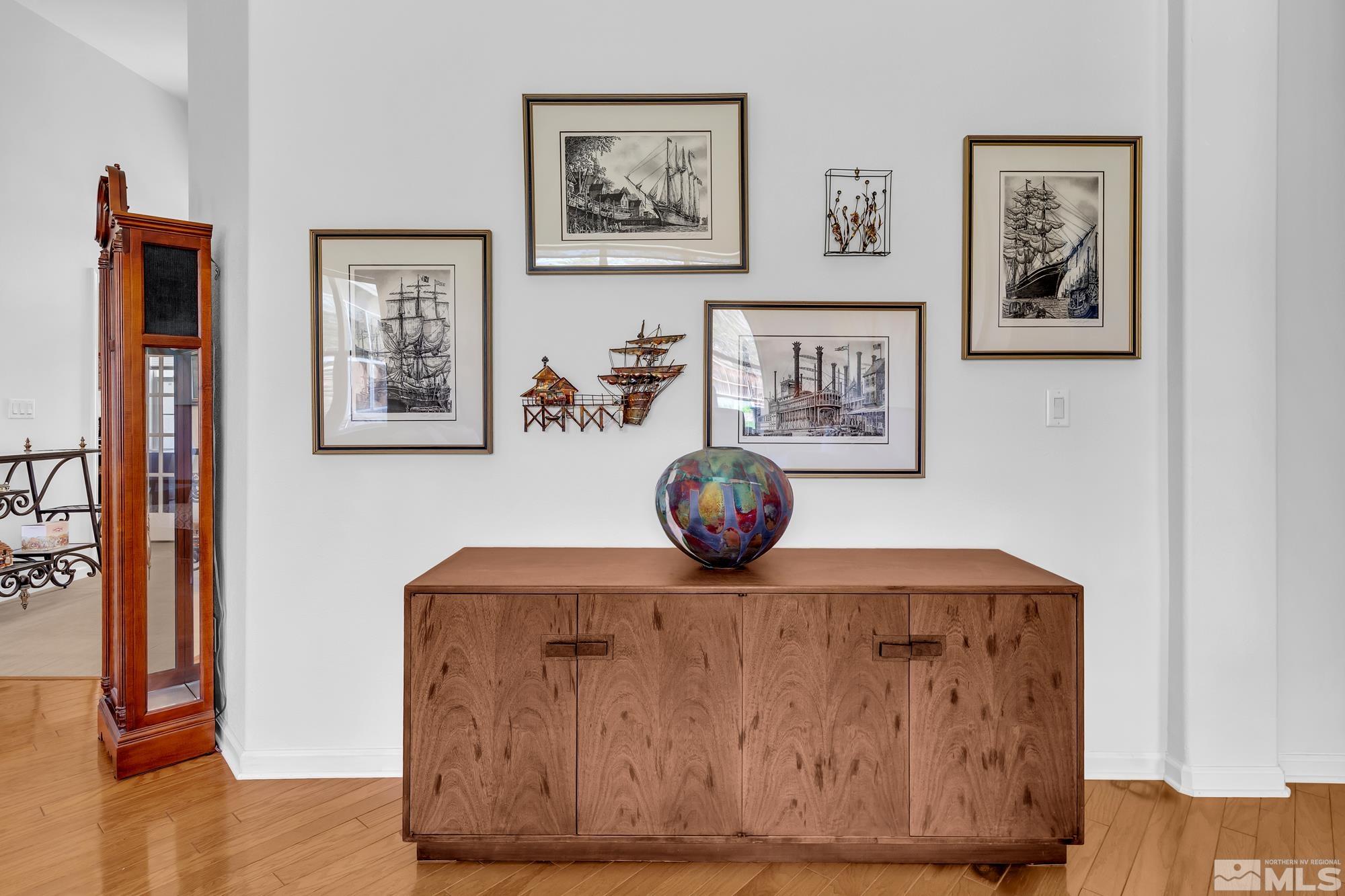 Bright interior with a wooden sideboard, colorful glass vessel, maritime prints, and tall wooden clock.