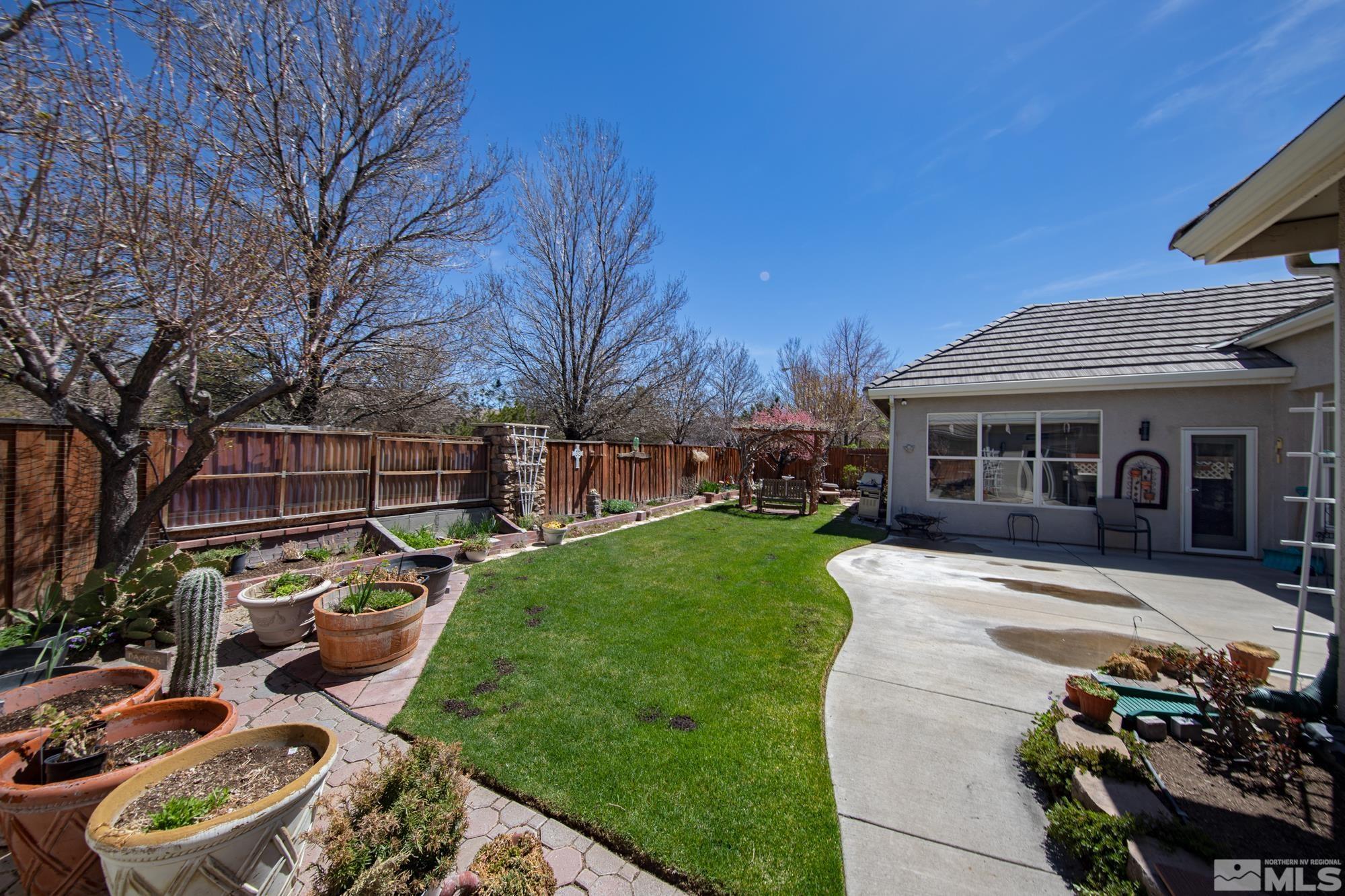 Fenced backyard with a green lawn, curved patio, and rows of potted plants and garden beds.