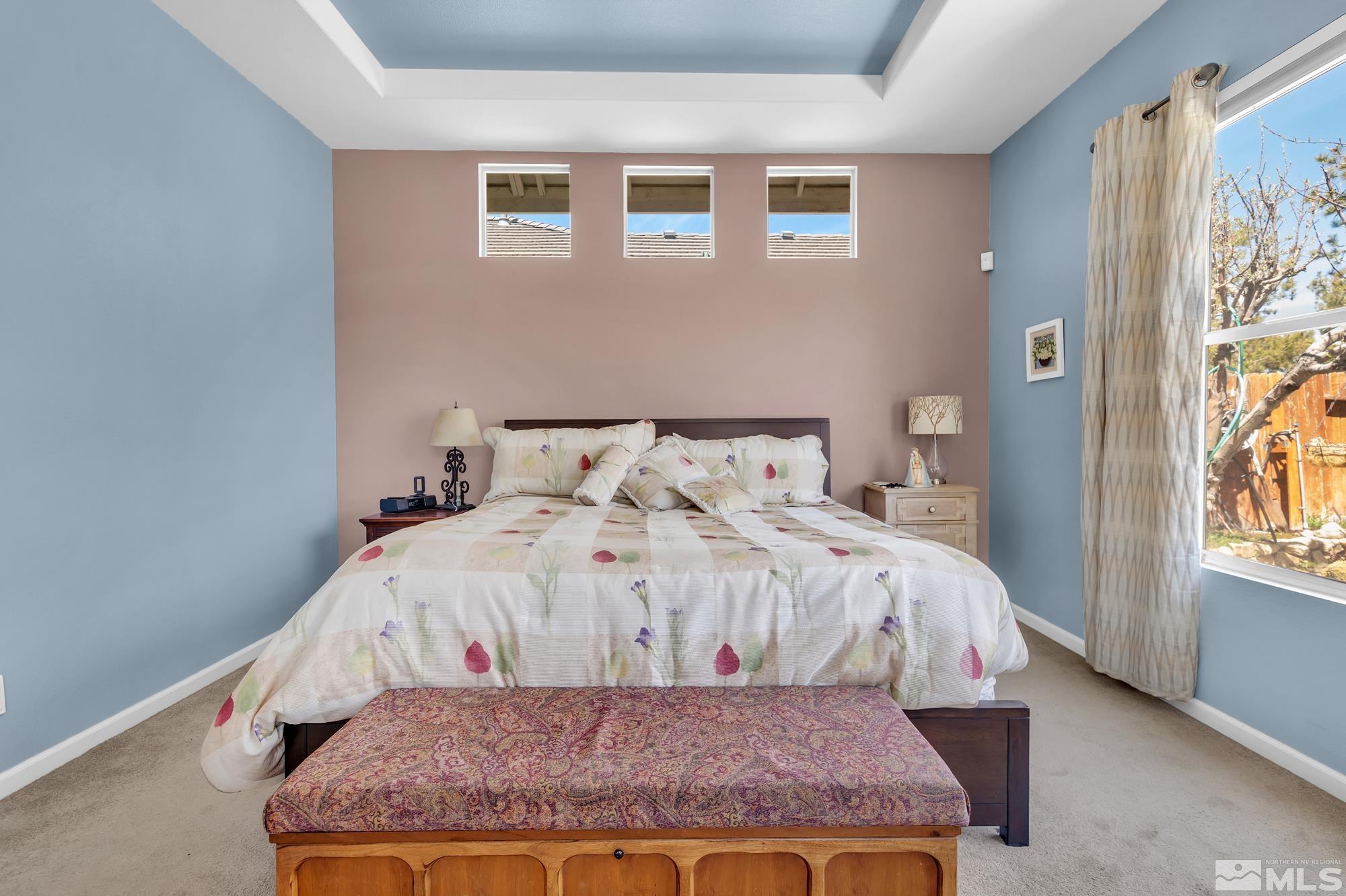 Furnished bedroom with a floral bedspread, three high windows, and a patterned bench at the foot of the bed.