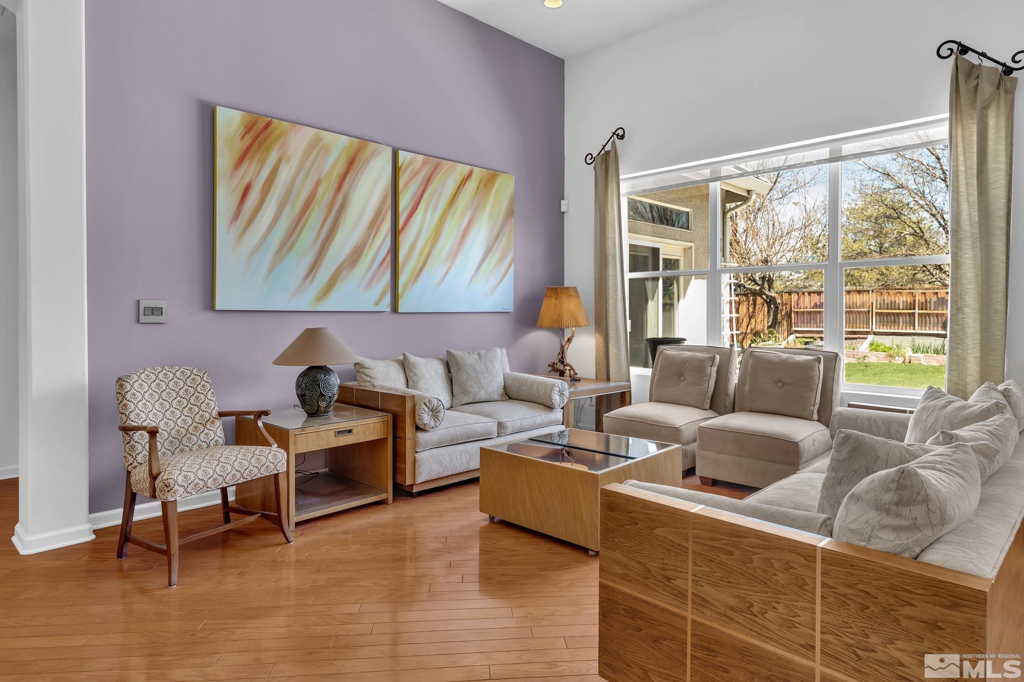 Living room with hardwood floors, lavender accent wall, and large windows overlooking a fenced backyard.