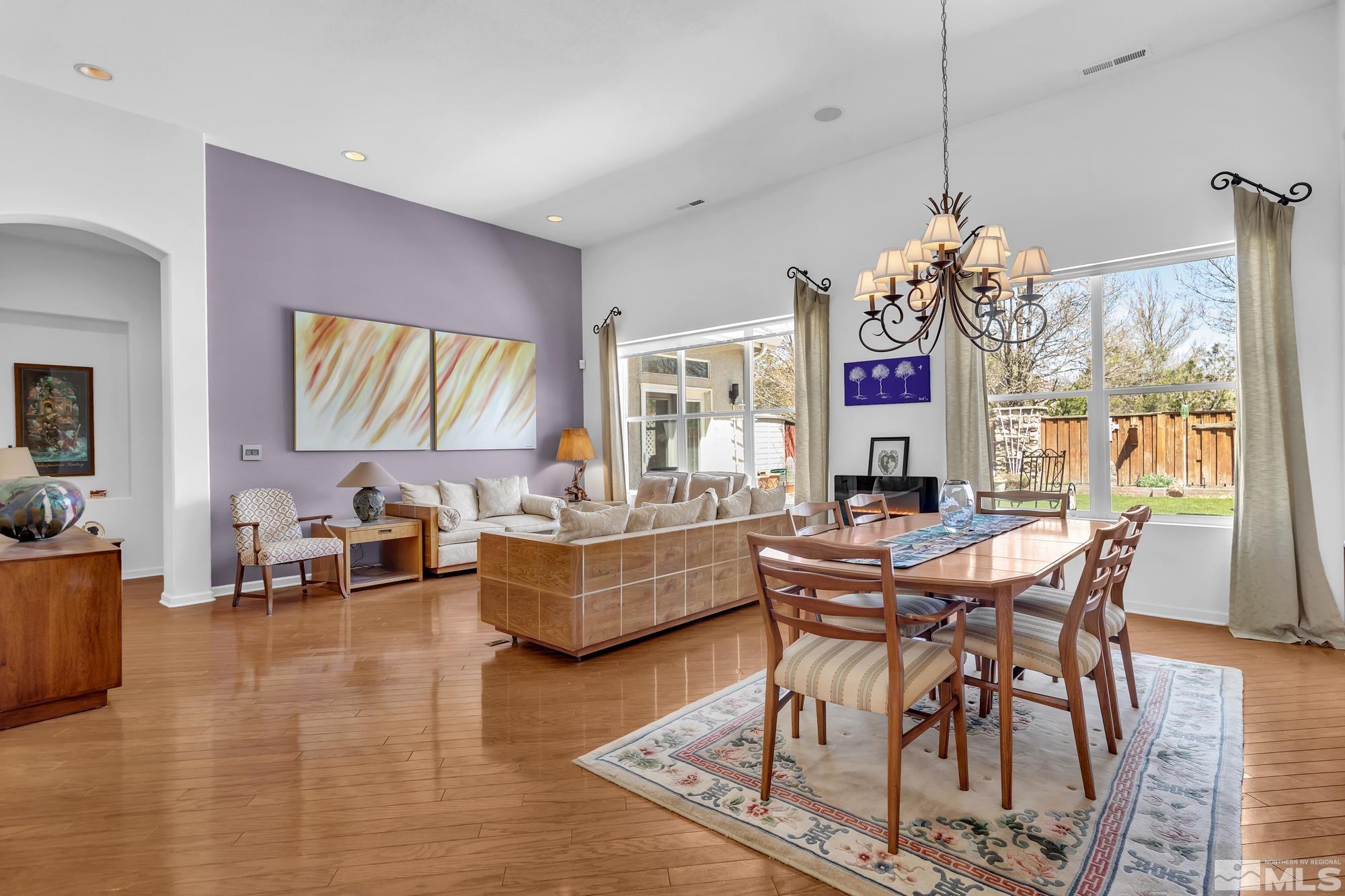 Open-plan living and dining room with hardwood floors, lavender accent wall, and large windows overlooking the backyard.