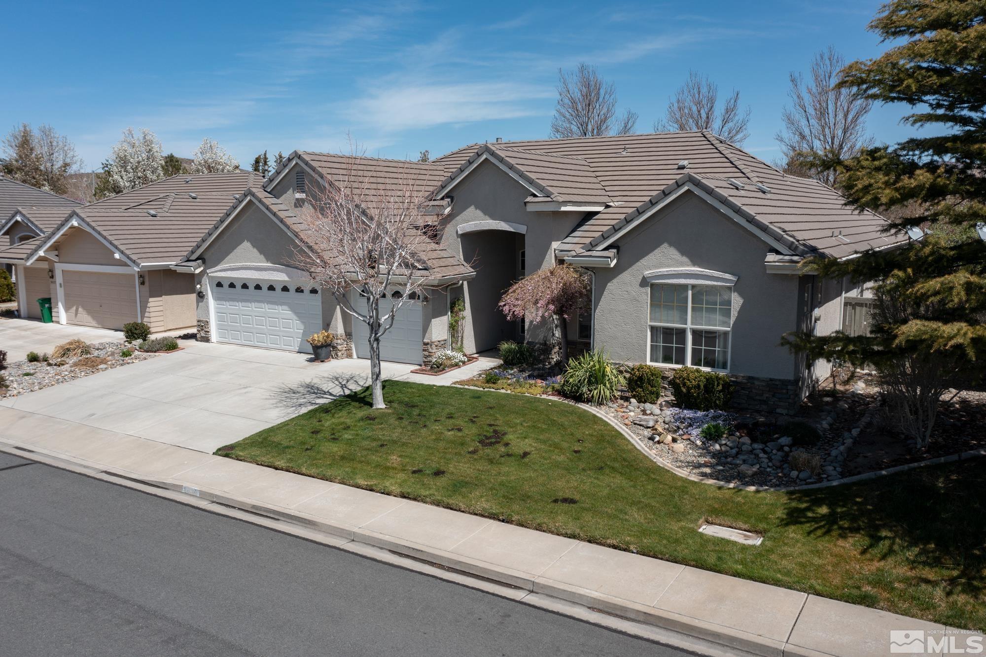 Gray stucco home with a tile roof, three-car garage, curved lawn, and landscaped rock beds.