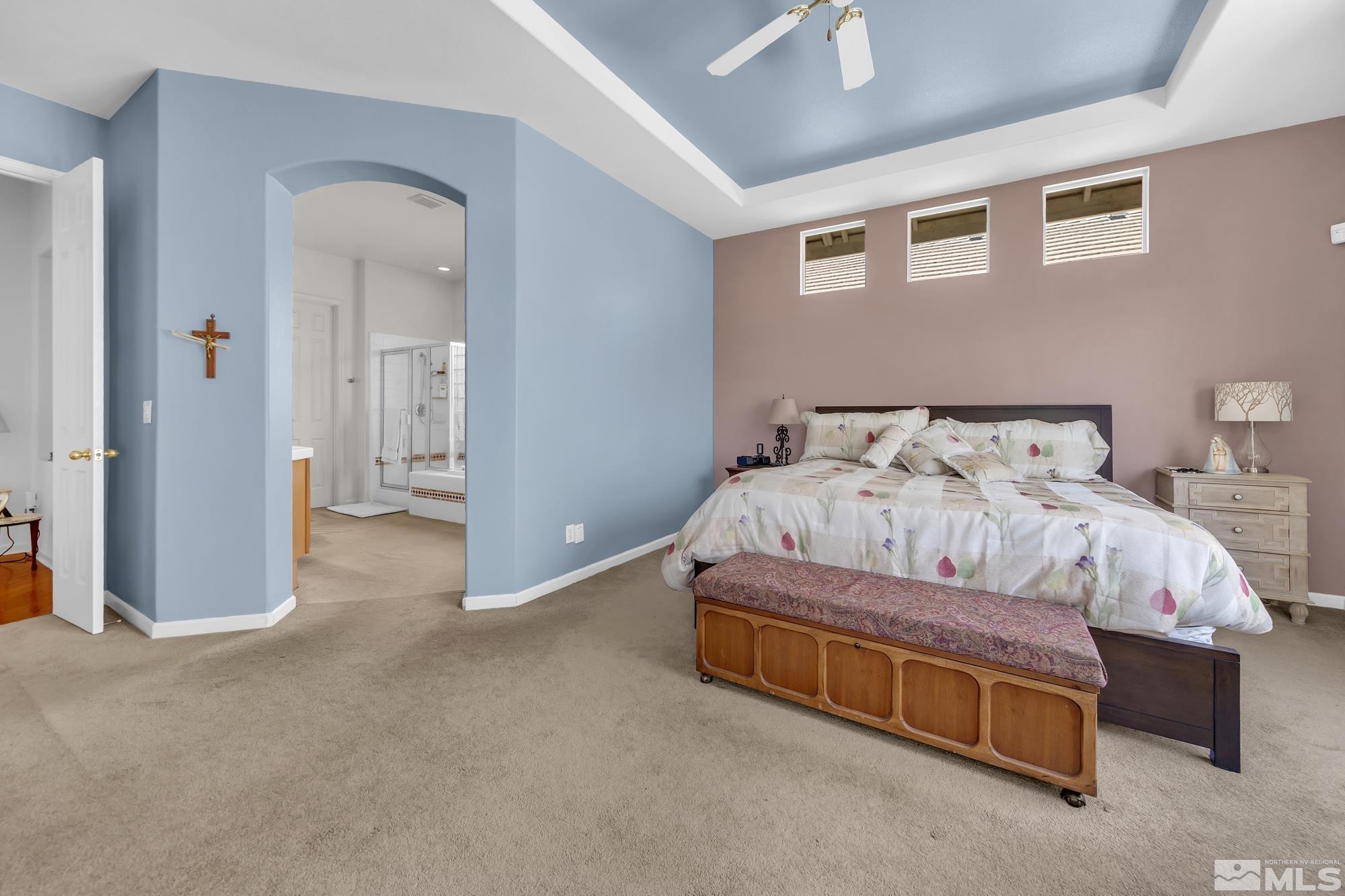 Bedroom with a blue tray ceiling, large bed, wooden bench, and arched opening to the ensuite bathroom.