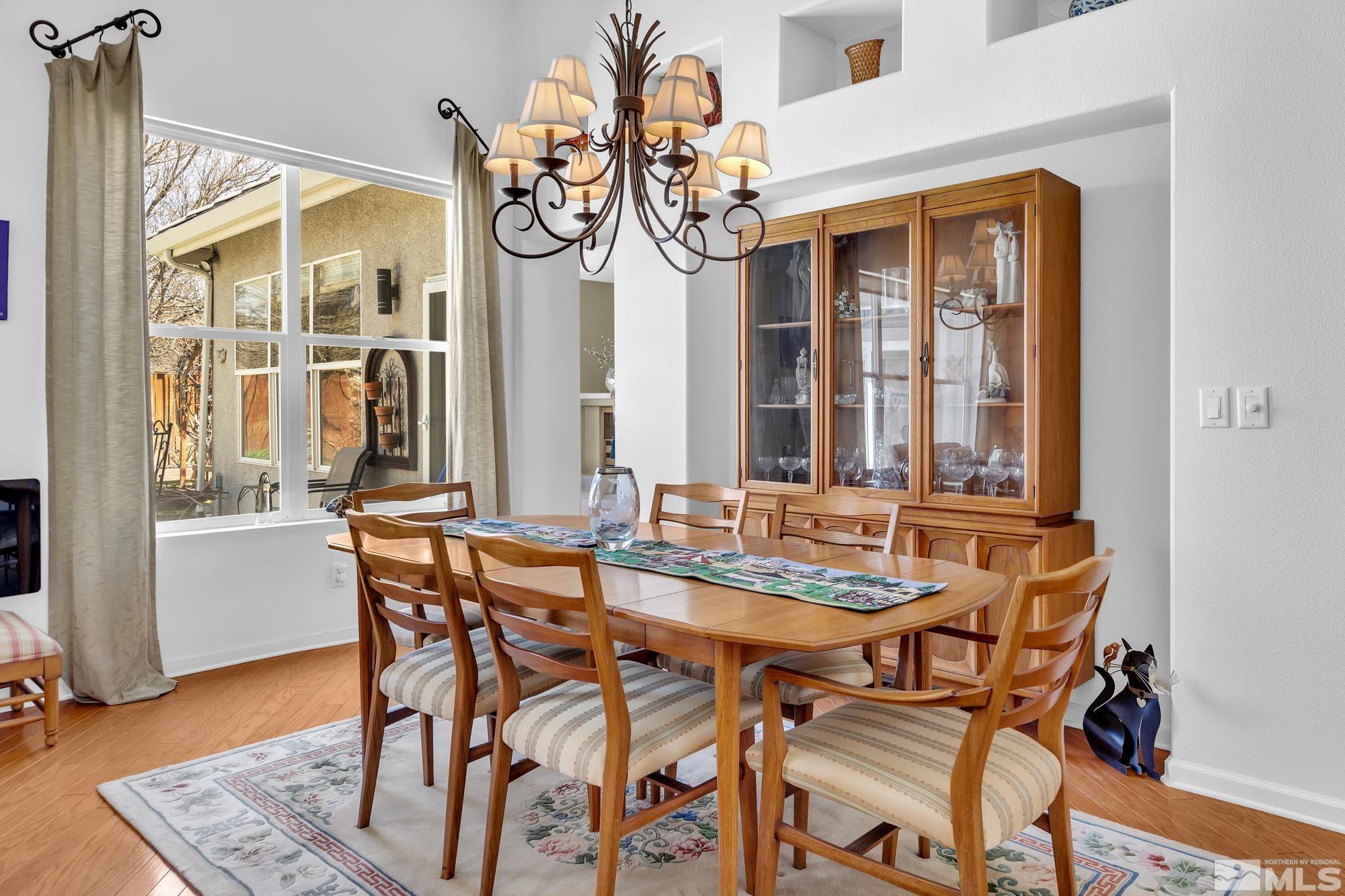 Dining room with a wooden table, glass-front china cabinet, chandelier, and large curtained windows.