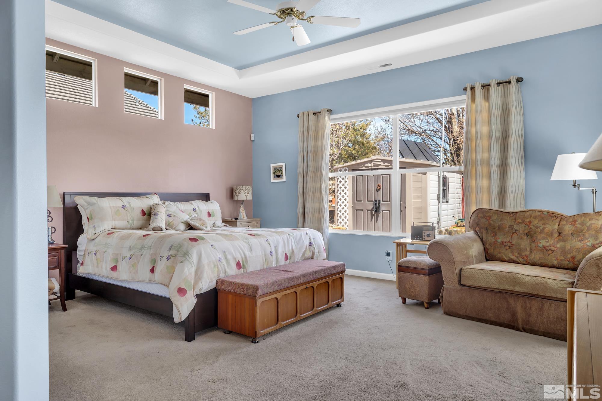 Furnished bedroom with floral bedding, a wood bench, and a wide window overlooking an outbuilding.