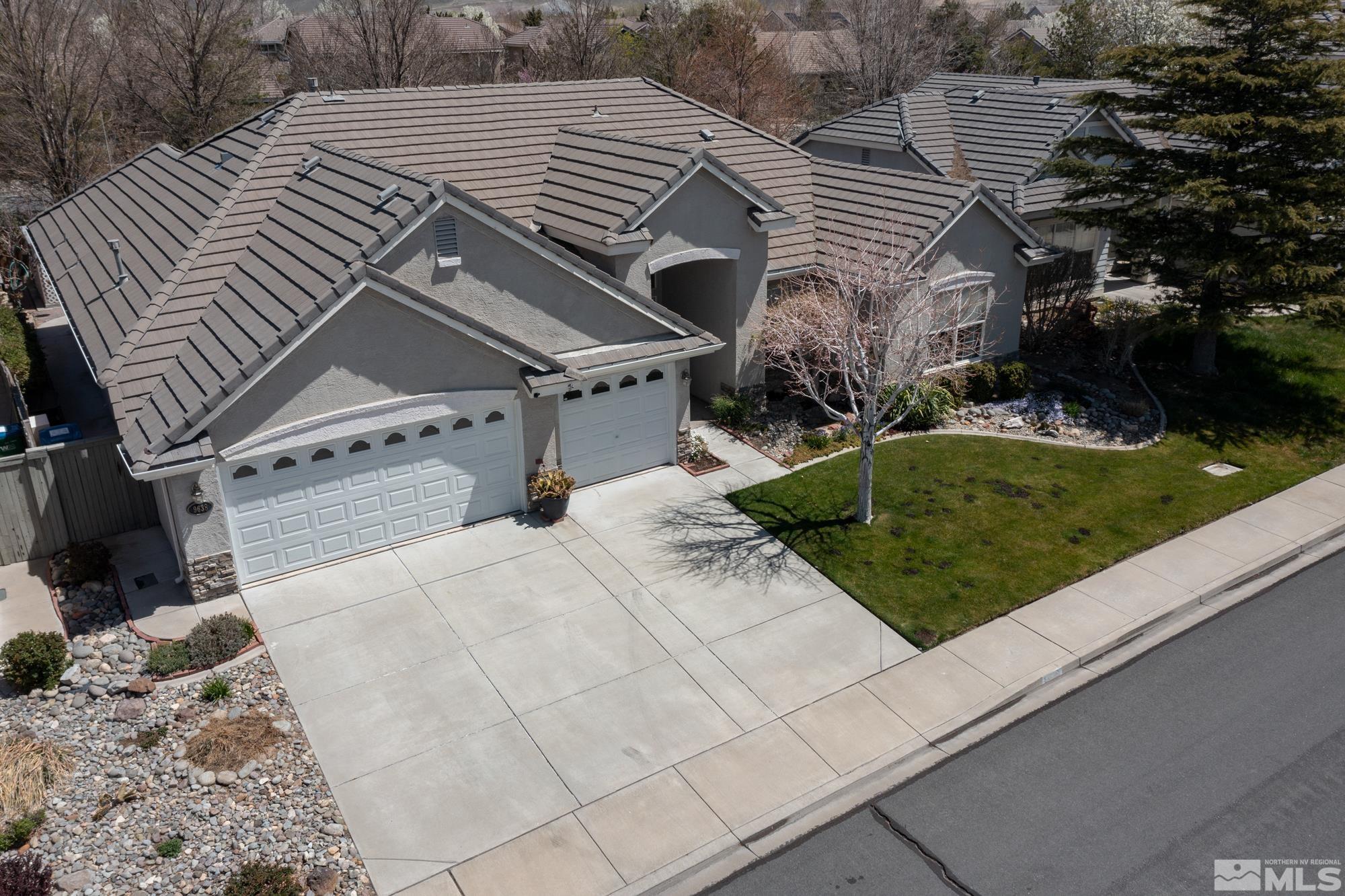 Aerial view of a gray stucco home with a multi-gabled roof, two-car garage, wide driveway, and front lawn.