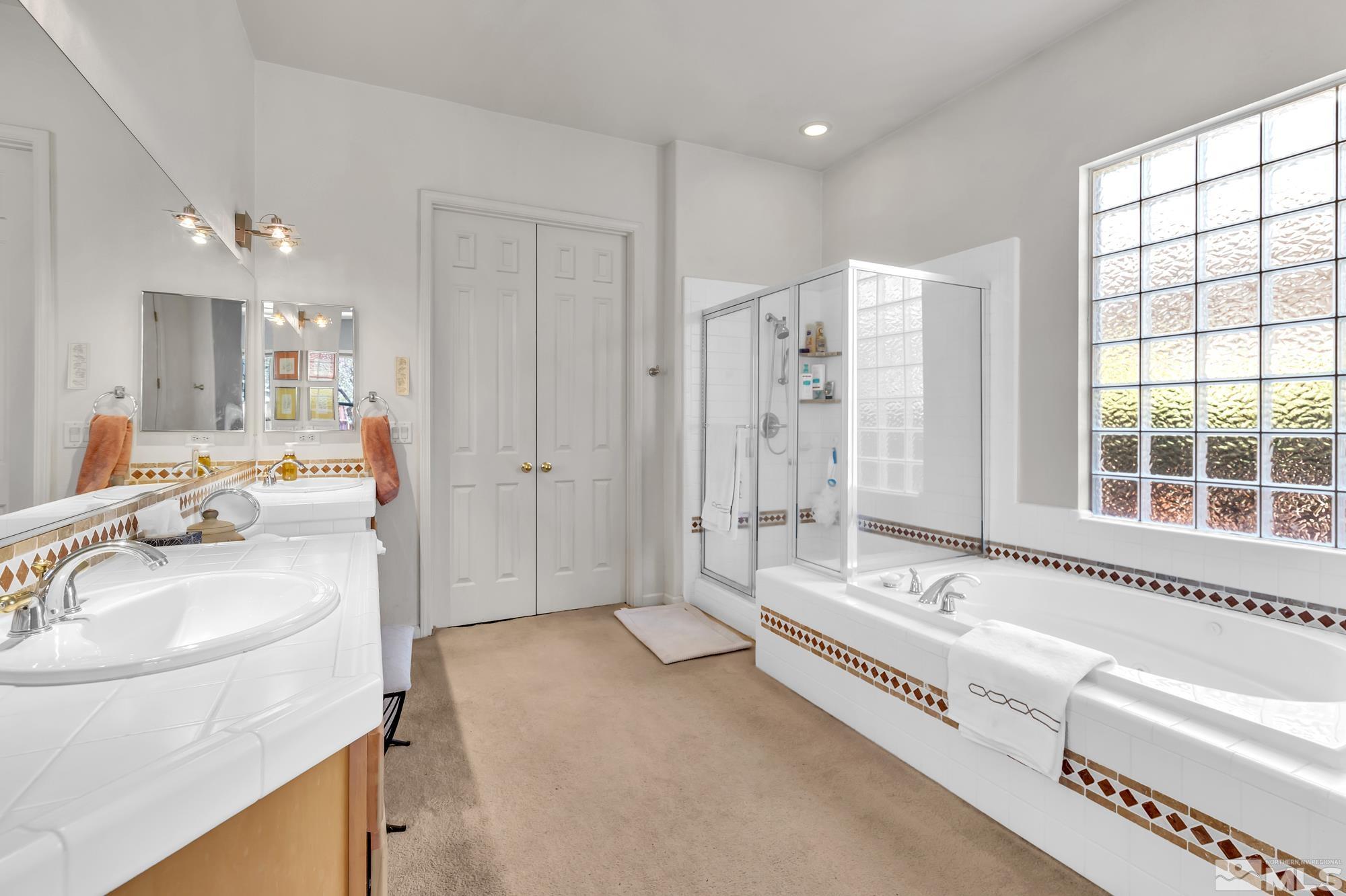 Bright bathroom with double-sink vanity, glass shower, and tub beneath a large glass-block window.