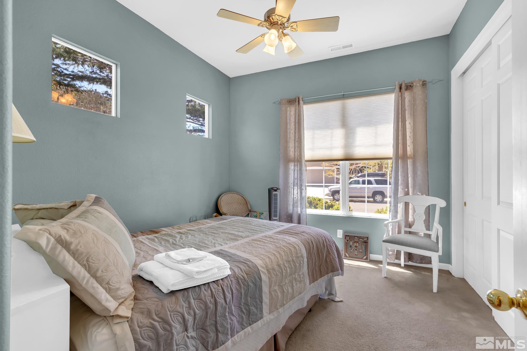 Furnished bedroom with a made bed, folded towels, blue-gray walls, and a large curtained window.