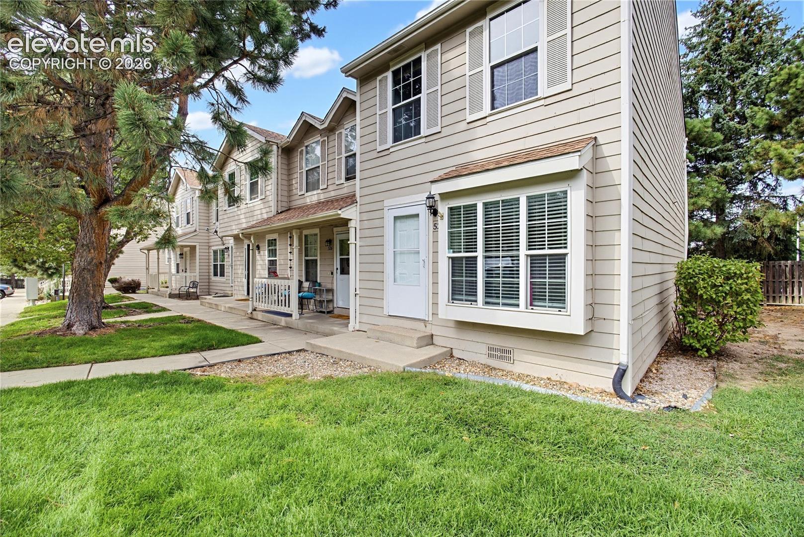 Beige two-story townhomes with covered porches beside a large lawn and mature pine trees.
