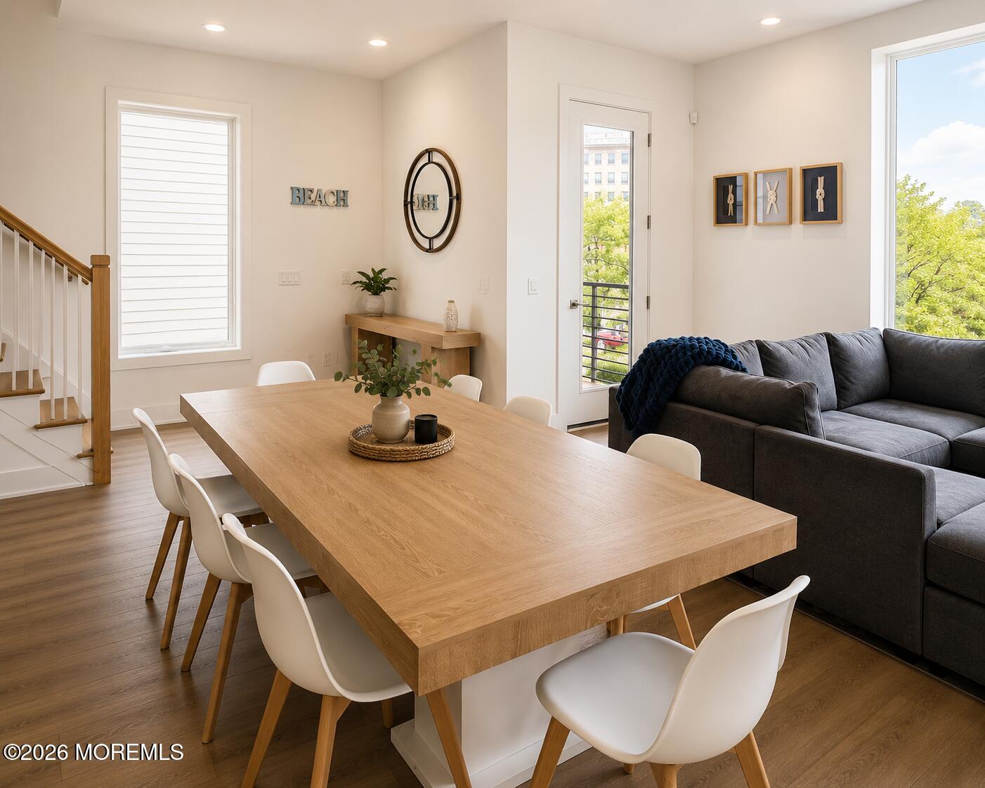 Bright office meeting area with a long wooden table, white chairs, gray sectional, and large windows.