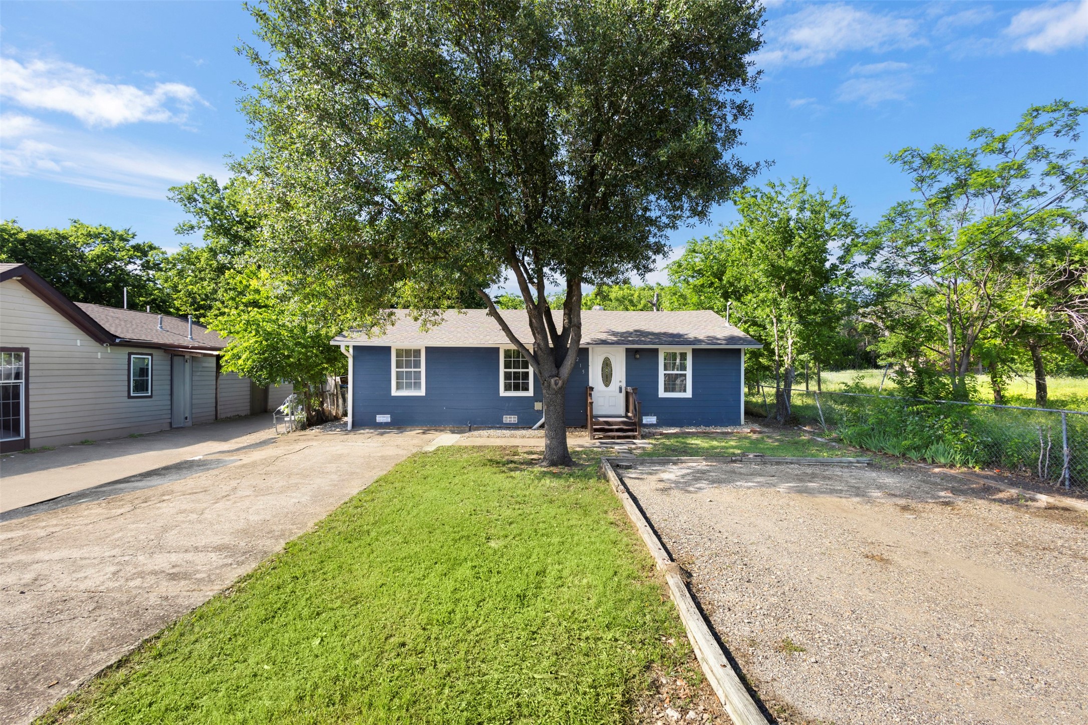 Blue single-story house beneath a large tree, with a concrete driveway and gravel parking area.