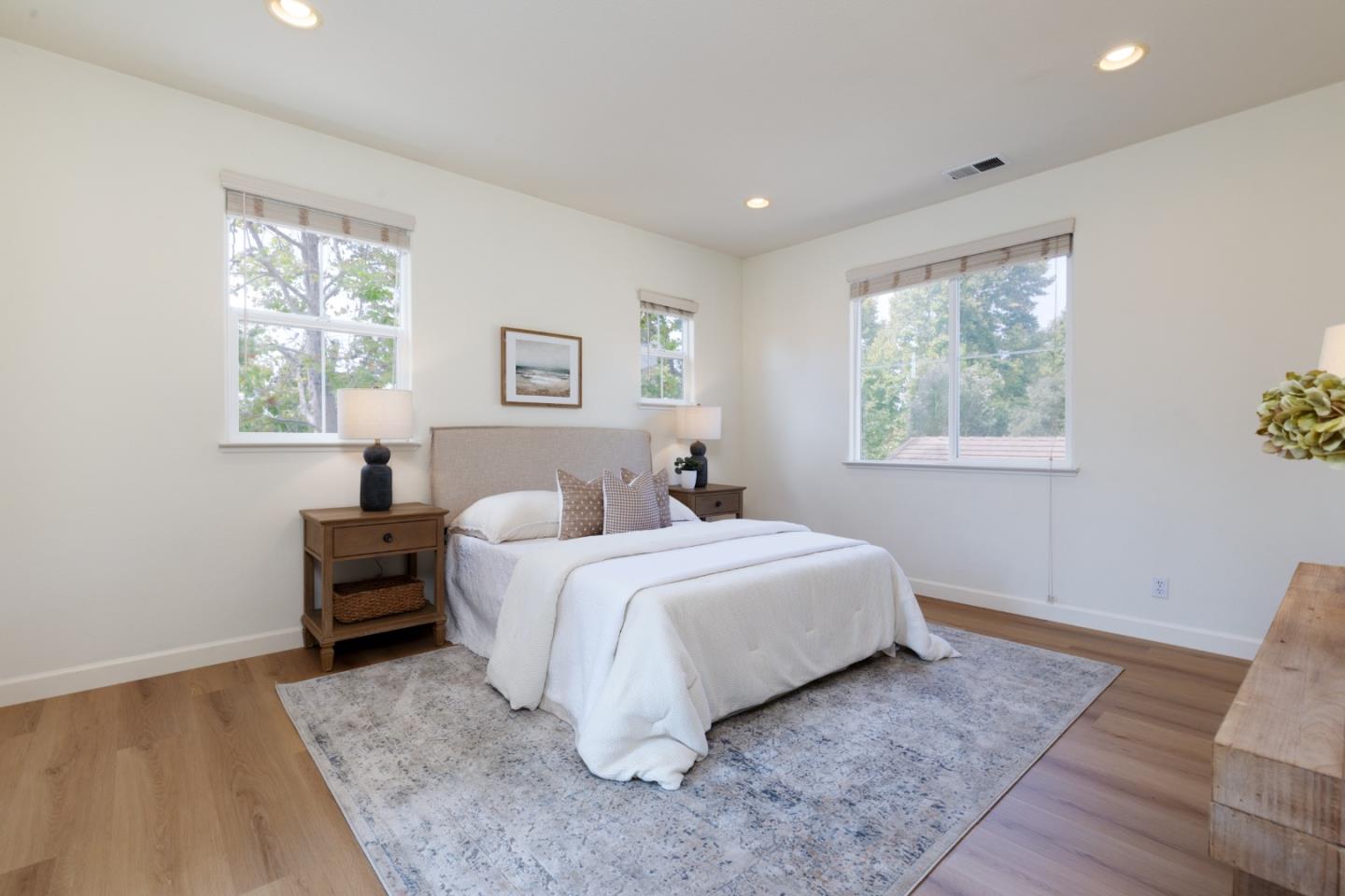Staged bedroom with a white bed, wood nightstands, and three windows overlooking trees.