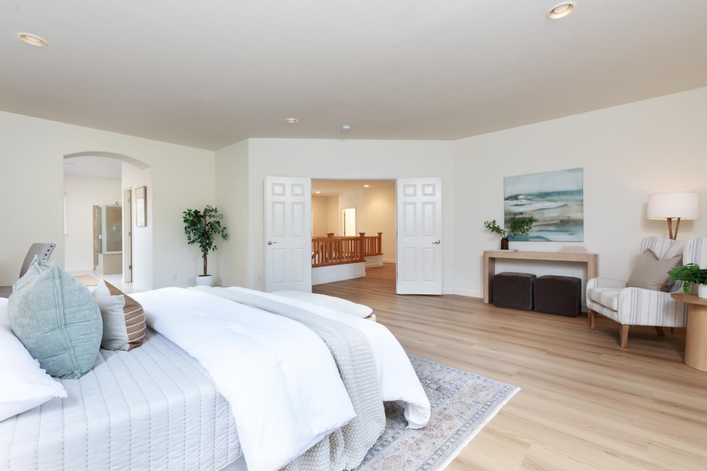 Staged bedroom with a white bed, light wood floors, and open double doors to the hallway.