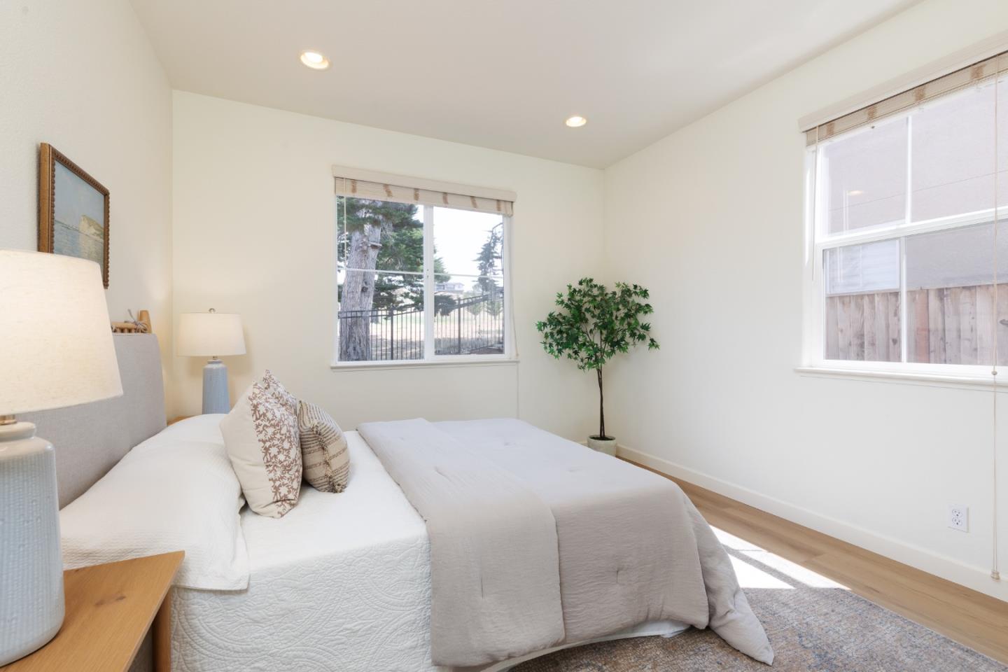 Bright staged bedroom with a large white bed, two windows, hardwood floors, and a potted plant.