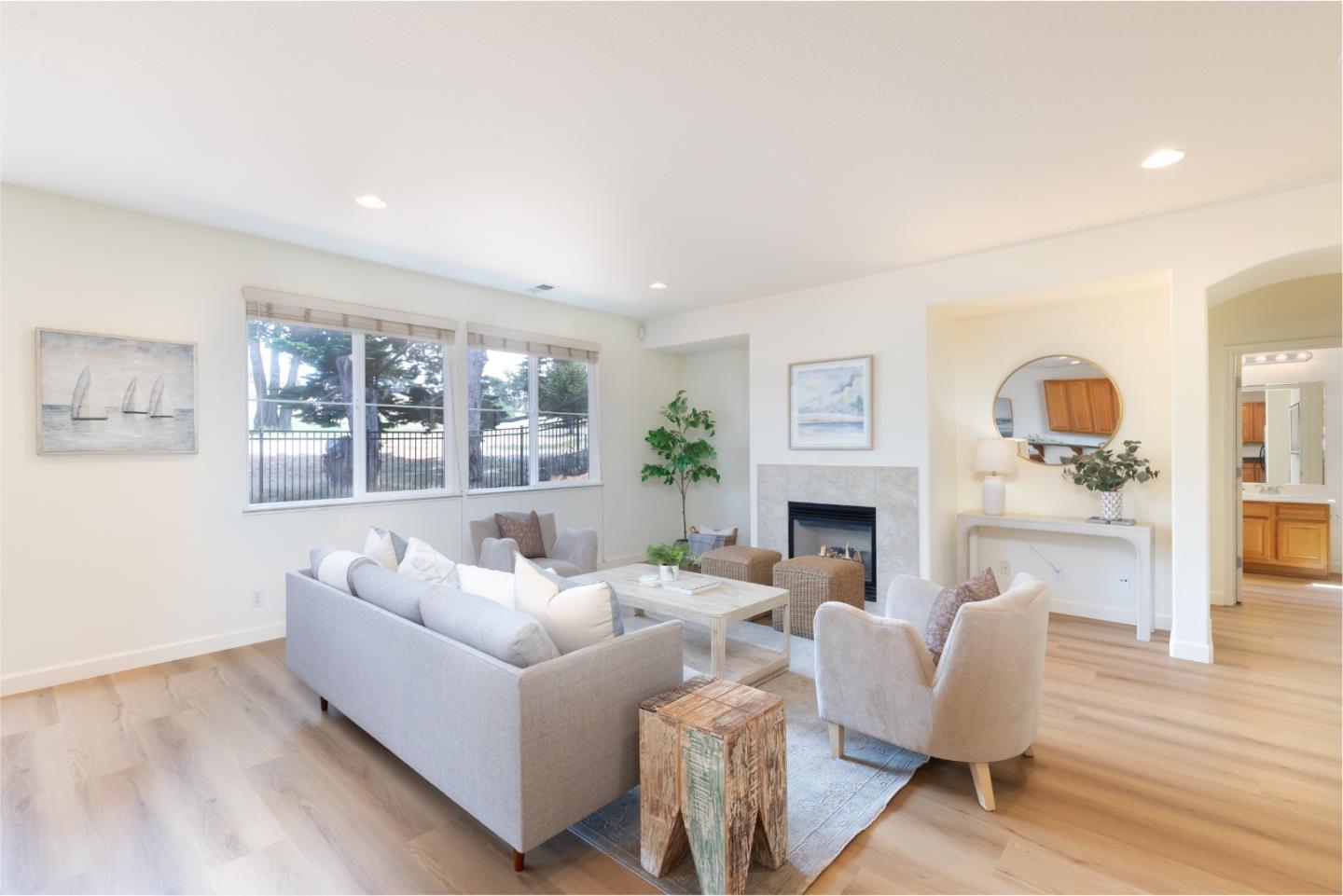 Furnished living room with a stone-surround fireplace, gray seating, and large windows overlooking trees.