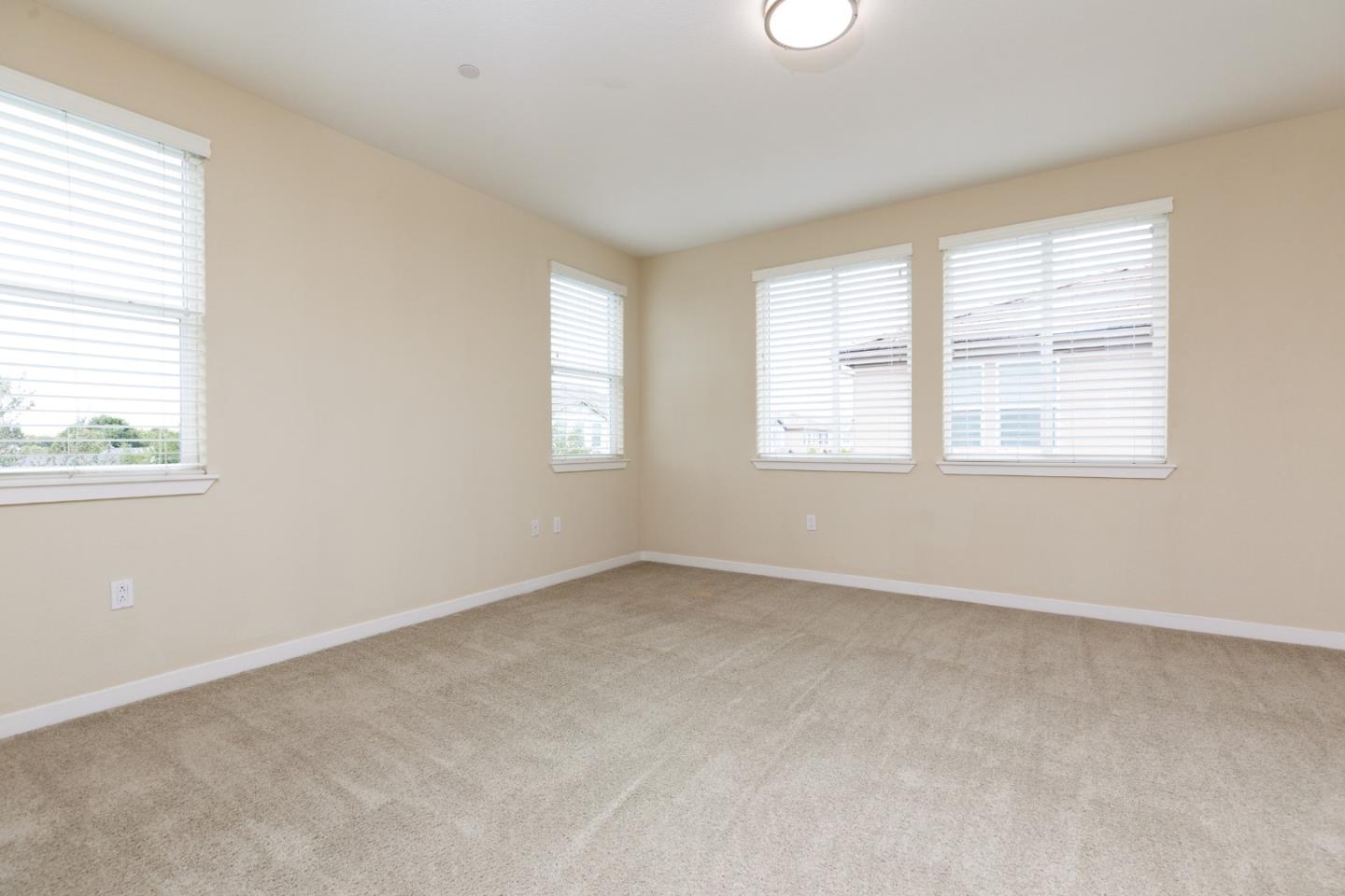 Empty carpeted room with beige walls, four windows with white blinds, and a ceiling light.