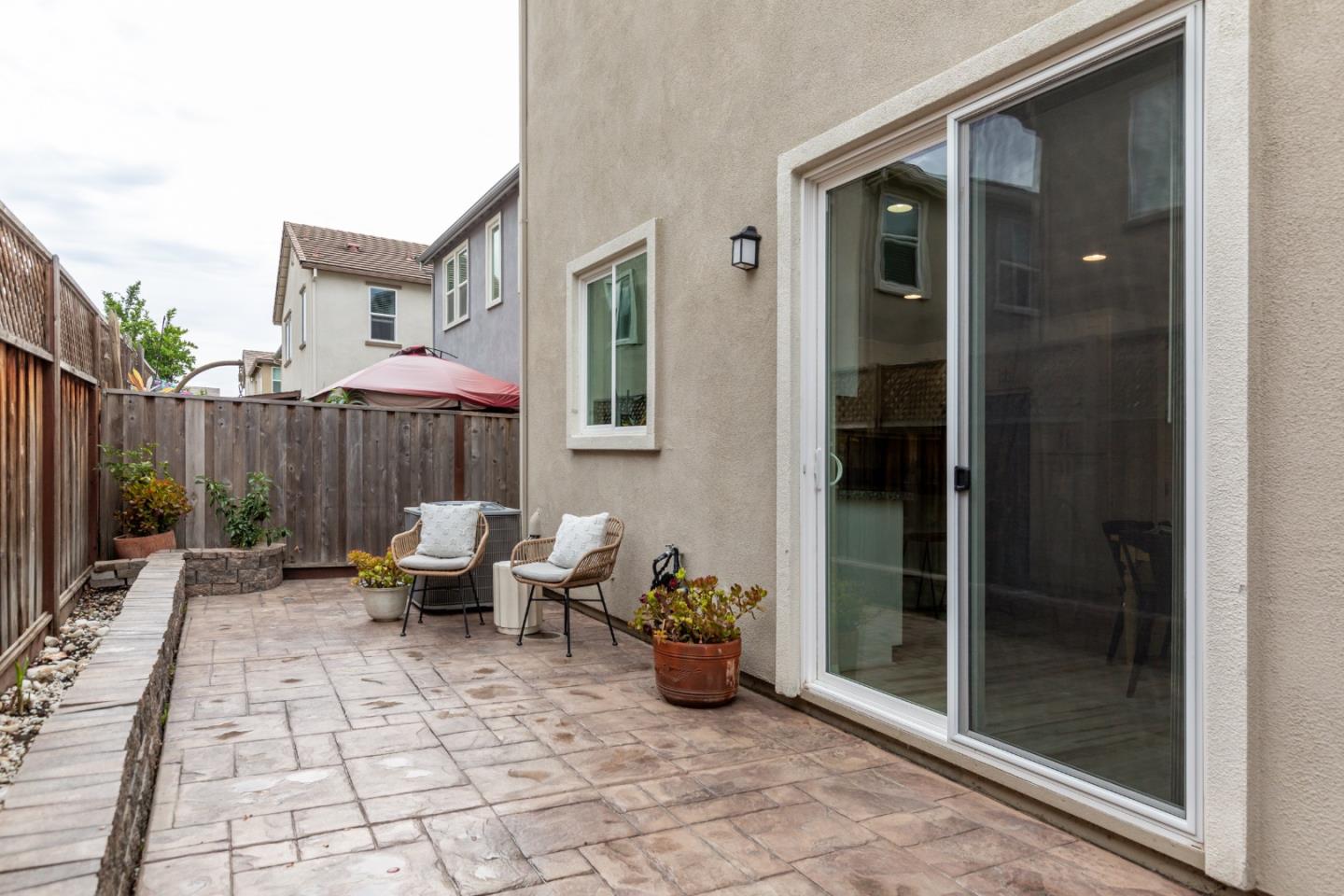 Backyard patio with stone paving, two cushioned chairs, potted plants, and a sliding glass door.
