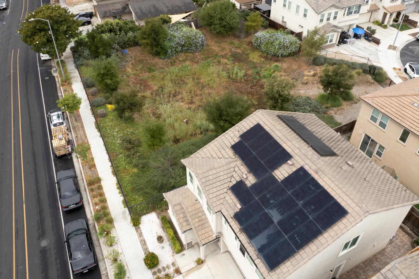Aerial view of a partly overgrown vacant lot beside homes and a residential street.