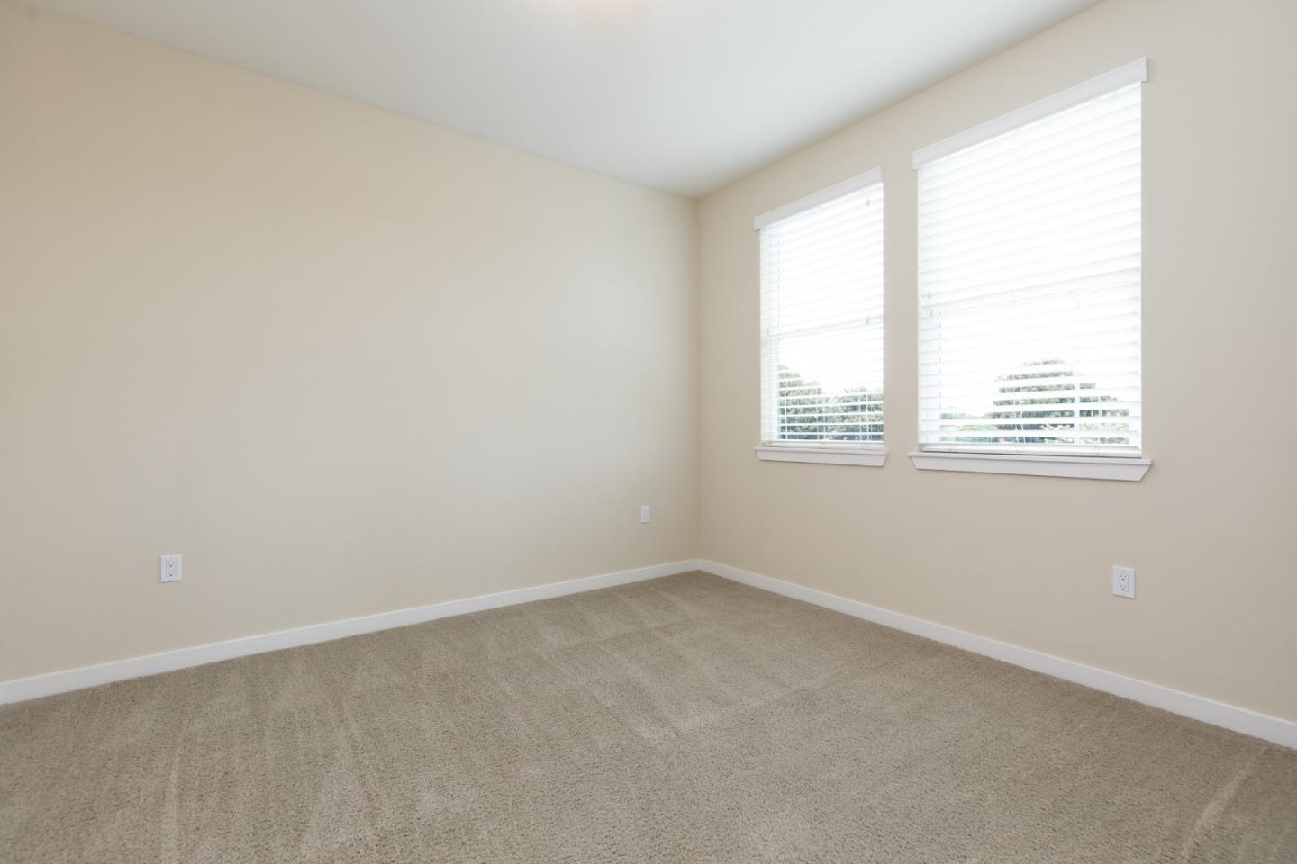 Empty bedroom with beige walls, taupe carpet, and two windows with white blinds.