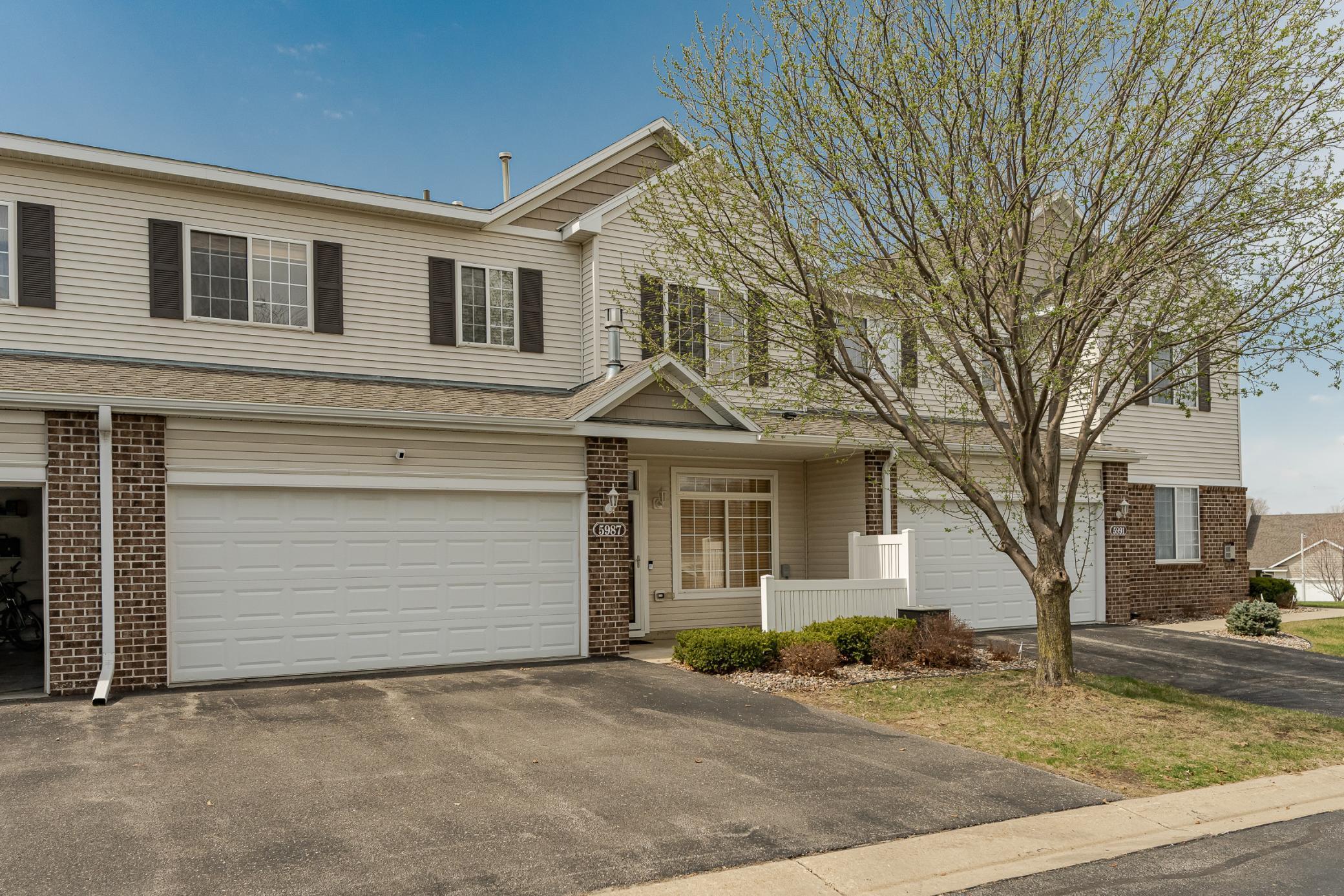 Two-story beige townhouse with brick accents, white garage doors, and a budding tree in front.