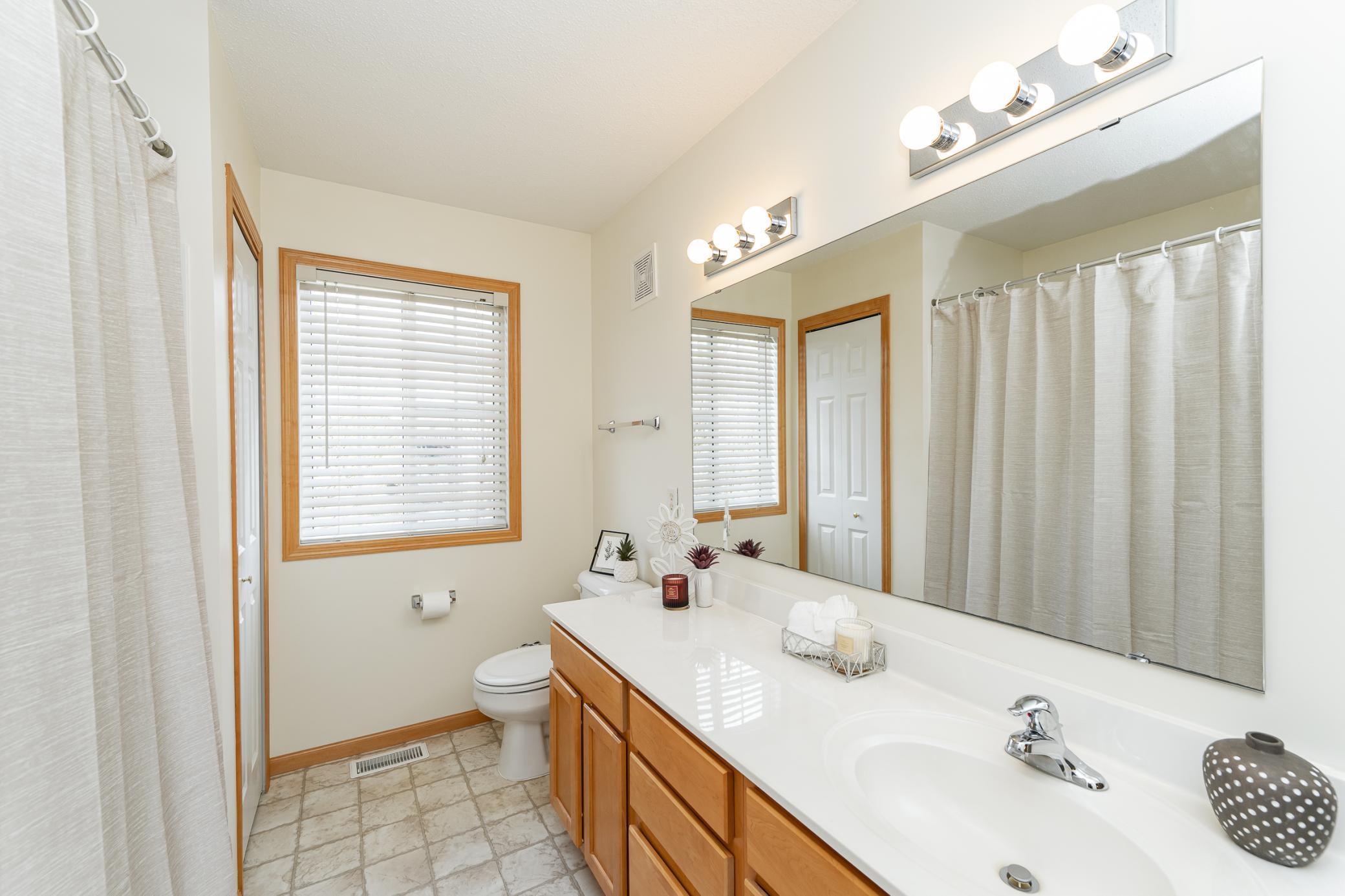 Bathroom with a long white vanity, wood cabinets, large mirror, and window with blinds.