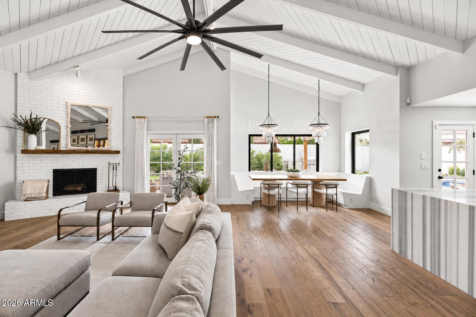 Open-plan living room with vaulted white-beamed ceiling, white brick fireplace, and adjoining dining area.