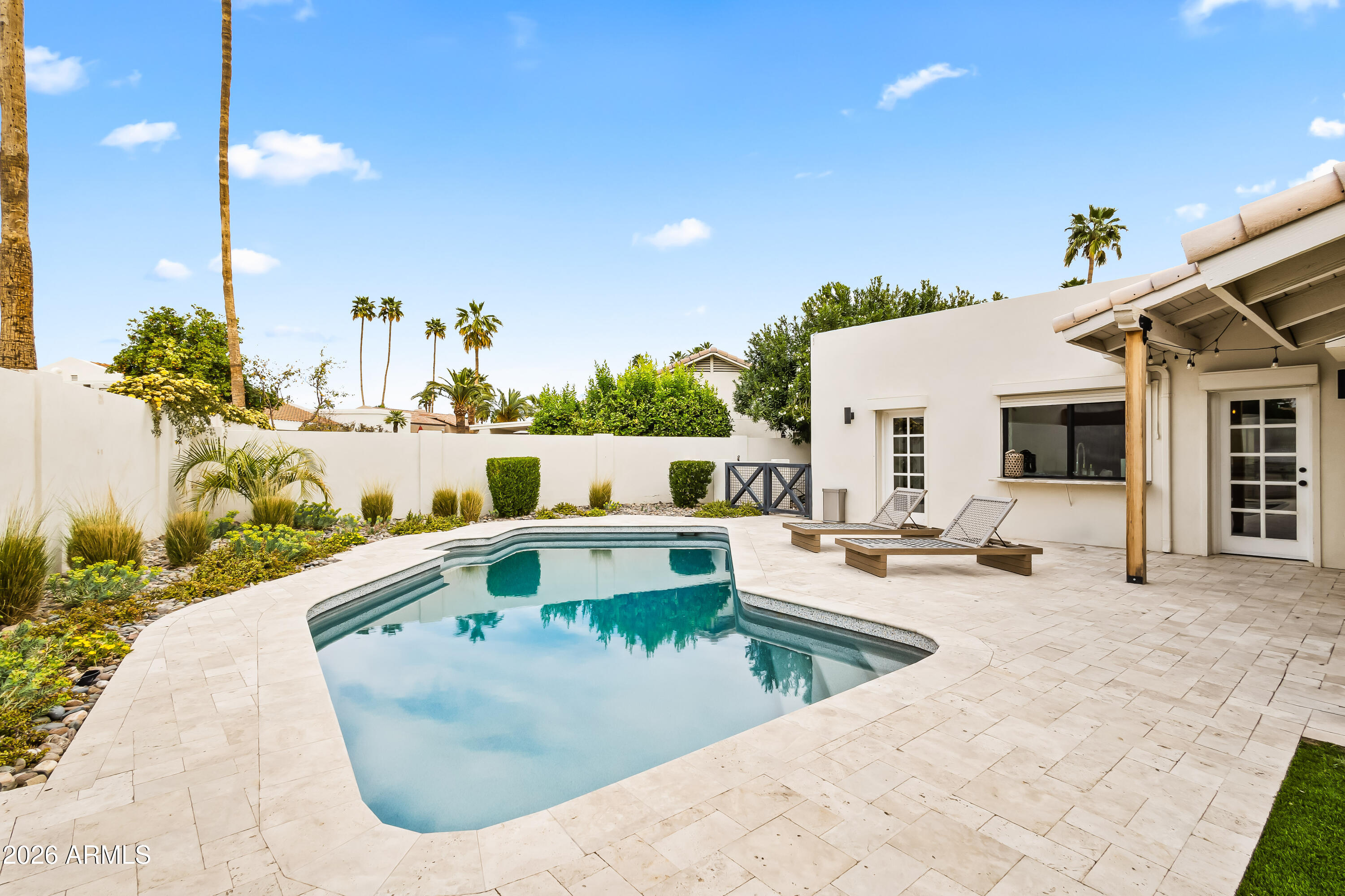 Backyard swimming pool with a pale stone patio, lounge chairs, and palm trees.
