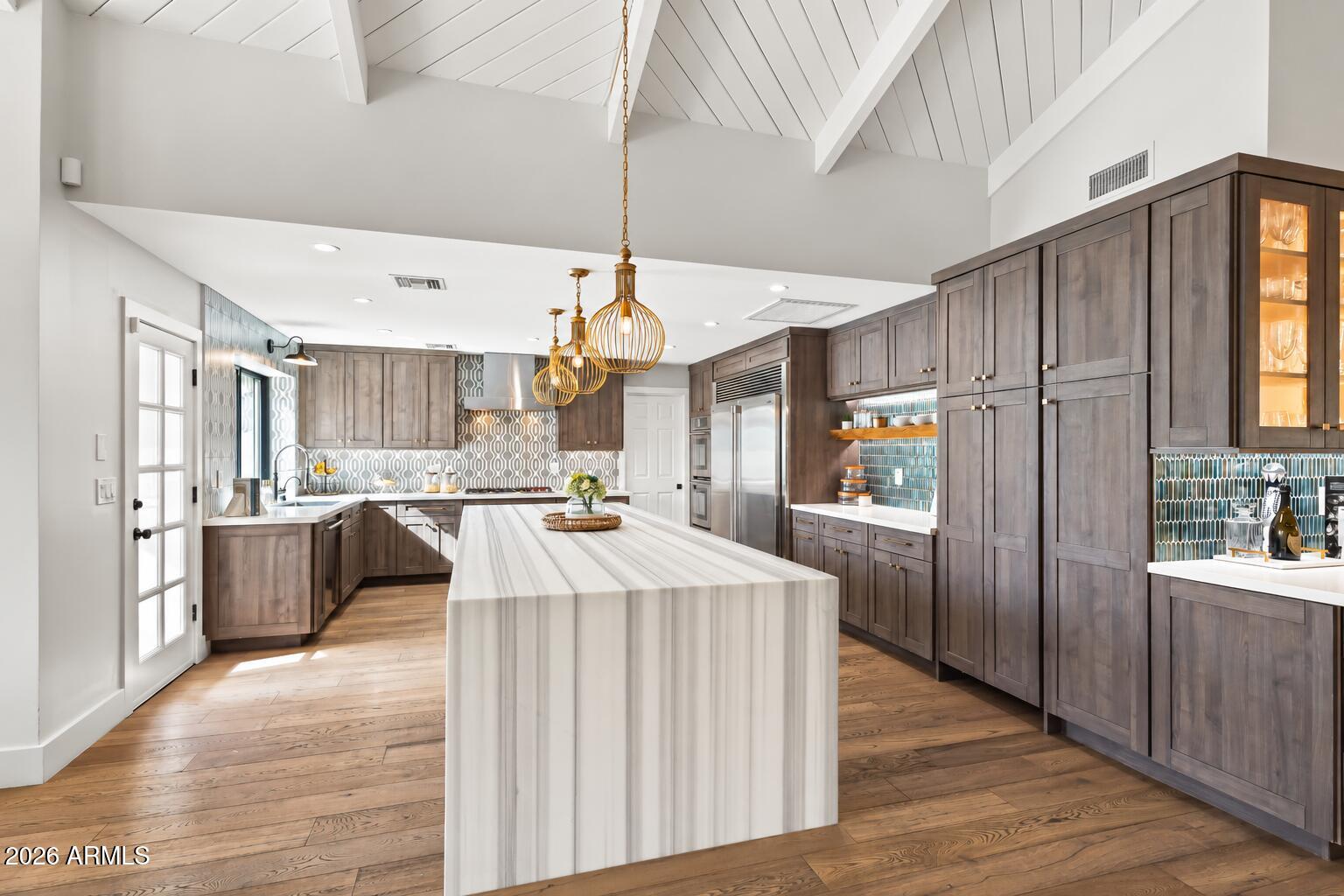 Kitchen with a large veined island, dark wood cabinets, teal tile backsplash, and vaulted beam ceiling.